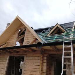 PATRYK FLORSZ - Budowa dachu drewnianego domu z widoczną konstrukcją więźby dachowej, deskowaniem, membraną dachową oraz pracownikiem na dachu i drabiną opartą o ścianę.