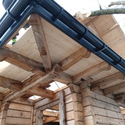 PATRYK FLORSZ - Wnętrze drewnianej konstrukcji dachu z widocznymi elementami więźby i zamontowanymi czarnymi rynnami, fragment drzewa w tle.