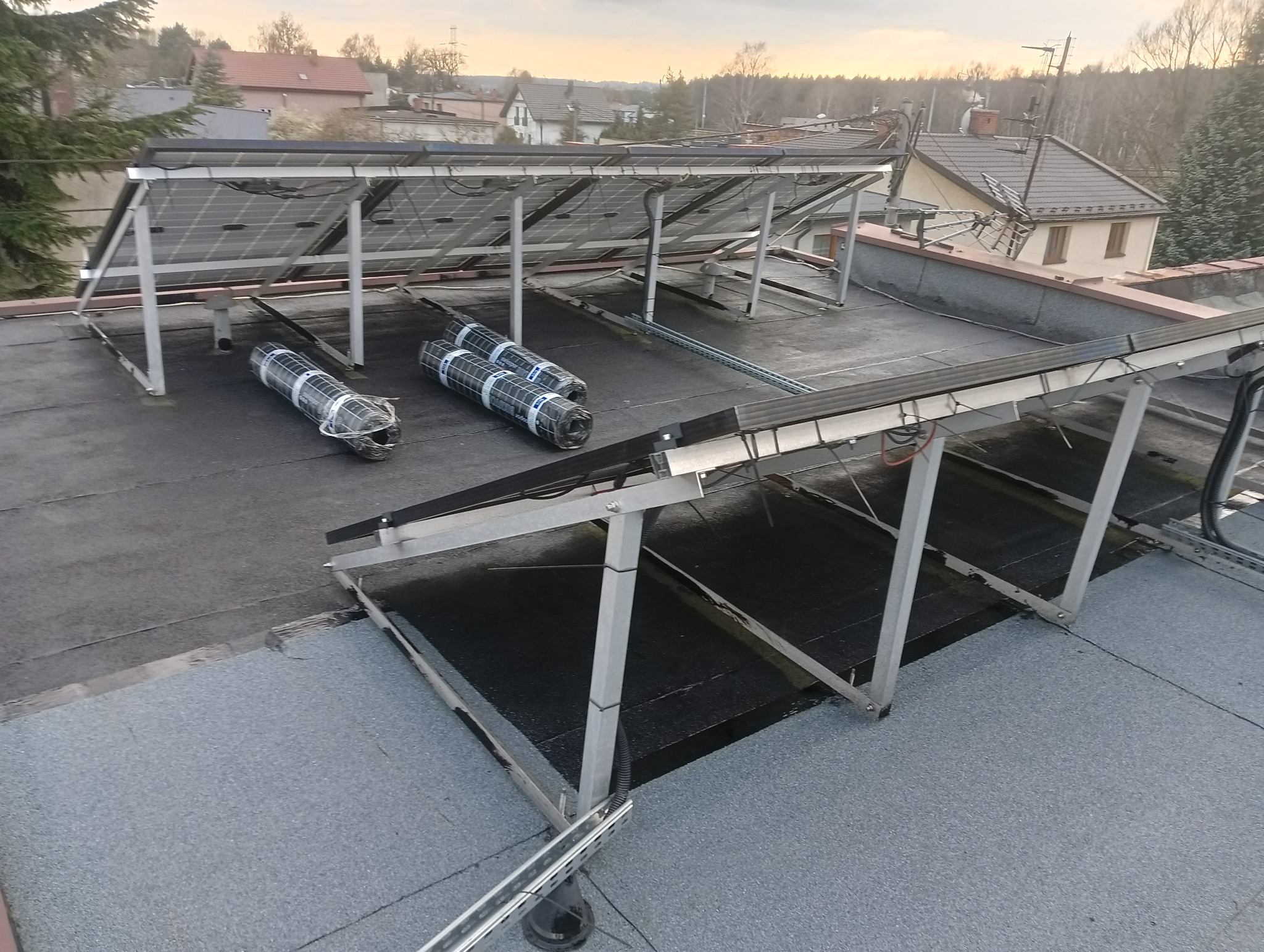 Płaski dach z zamontowaną konstrukcją pod panele słoneczne, trzy rolki papy bitumicznej w folii oczekują na rozłożenie, w tle zabudowa jednorodzinna.