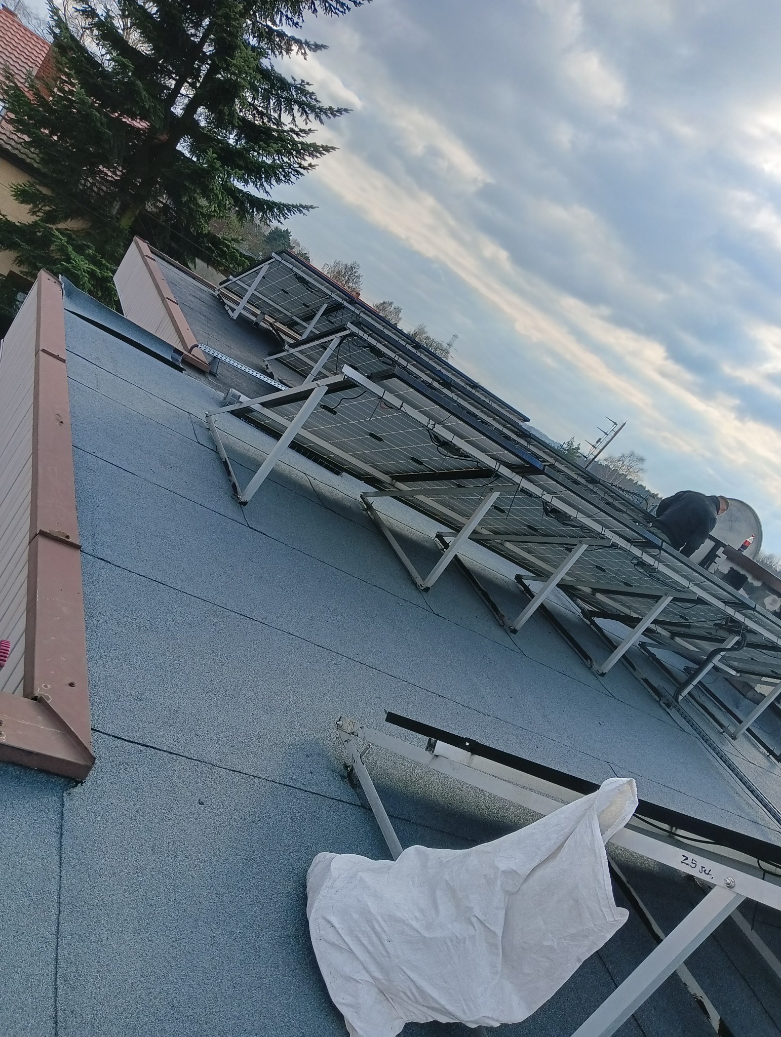 Montaż paneli fotowoltaicznych na dachu pokrytym papą, widoczny pracownik w ciemnej odzieży, białe płótno ochronne i metalowe konstrukcje wsporcze.