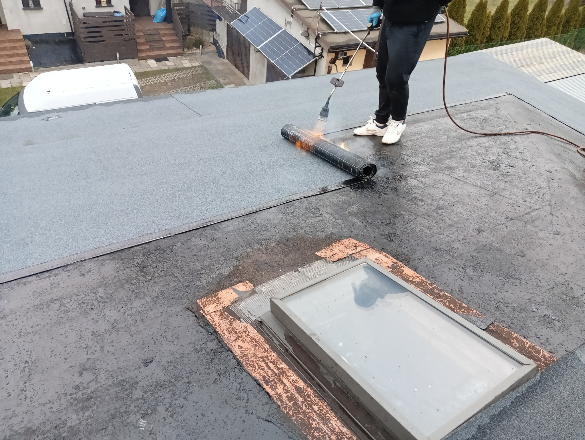 Pracownik w białych butach łączy papę termozgrzewalną z dachem płaskim za pomocą palnika, widoczny świetlik dachowy i domy w tle.