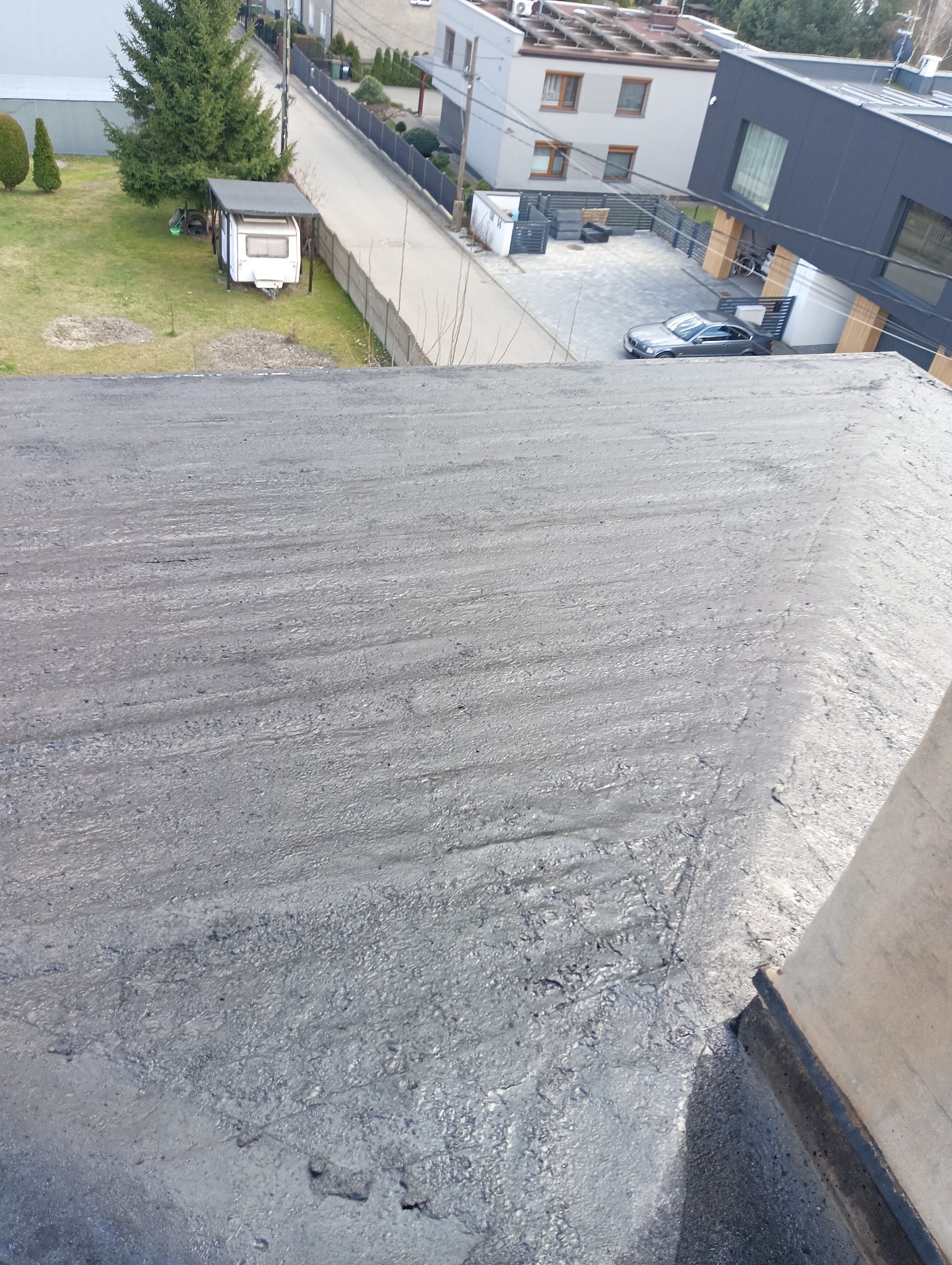 Płaski dach pokryty szarą, bitumiczną masą uszczelniającą, widok z góry na okoliczne budynki i ulicę.