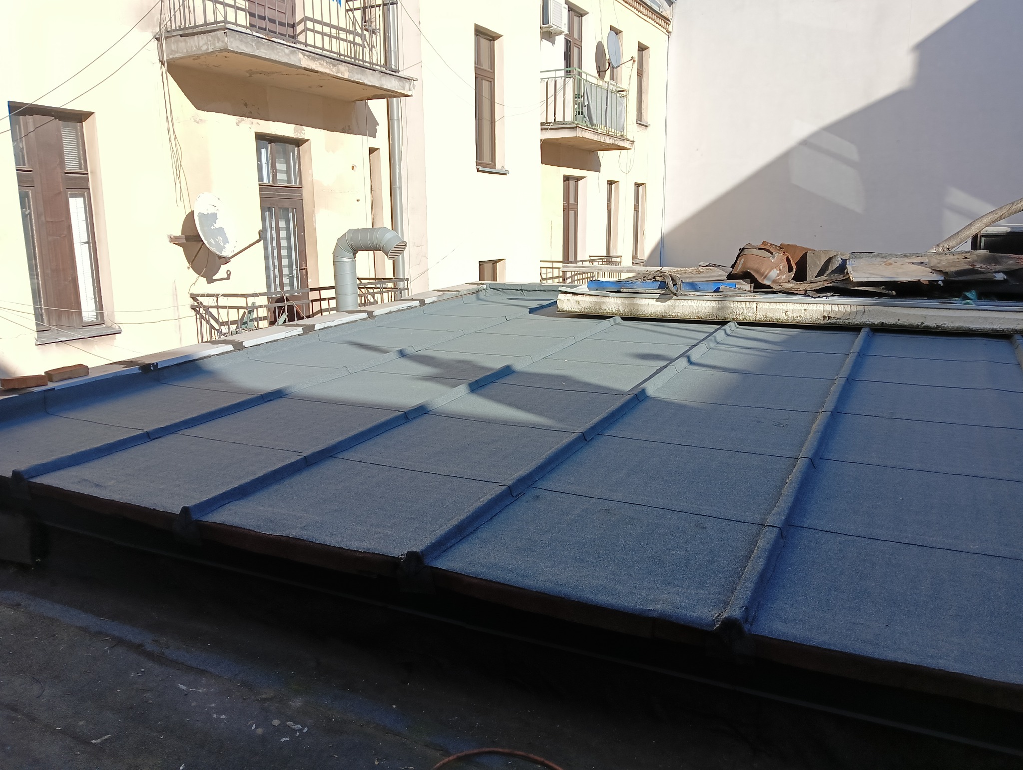 Płaski dach pokryty papą termozgrzewalną na budynku wielorodzinnym, widoczne detale łączeń i fragmenty konstrukcji dachu, w tle budynek z balkonami i instalacjami.