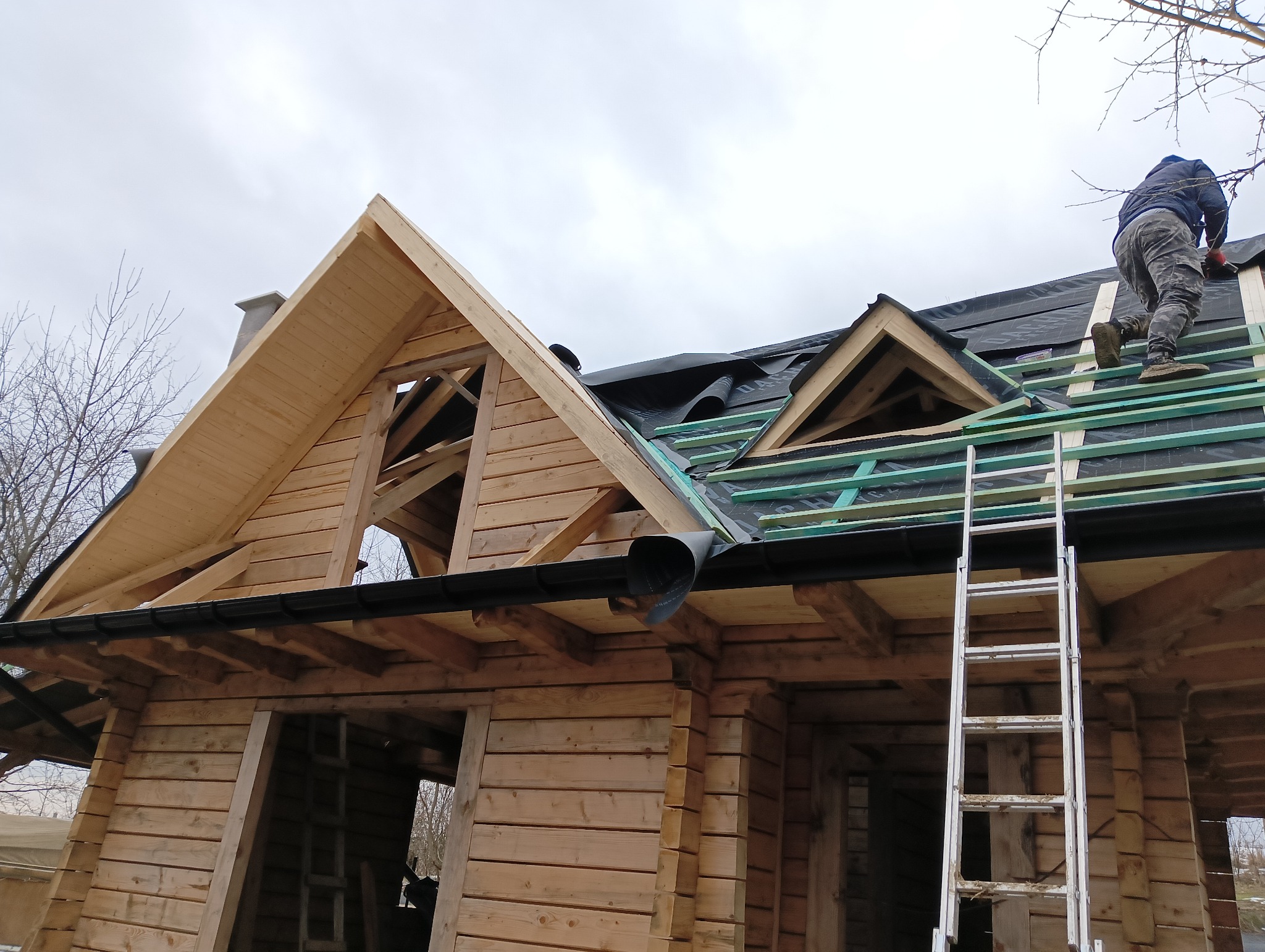 Budowa dachu drewnianego domu z widoczną konstrukcją więźby dachowej, deskowaniem, membraną dachową oraz pracownikiem na dachu i drabiną opartą o ścianę.