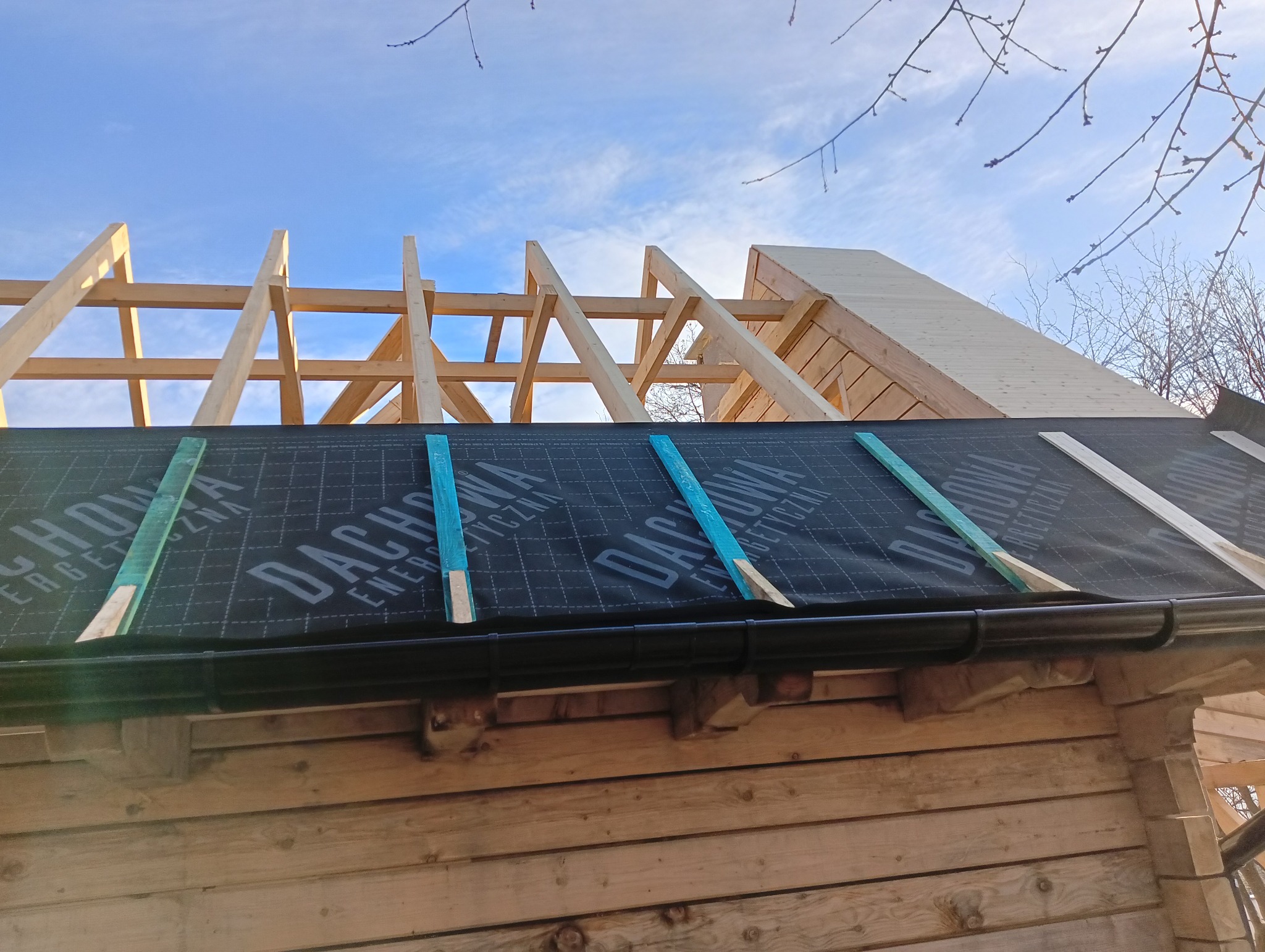Wiązar dachowy z drewnianych belek na budynku z bali, pokryty czarną membraną dachową z niebieskimi paskami mocującymi i zamontowaną rynną w kolorze czarnym, widoczne błękitne niebo.