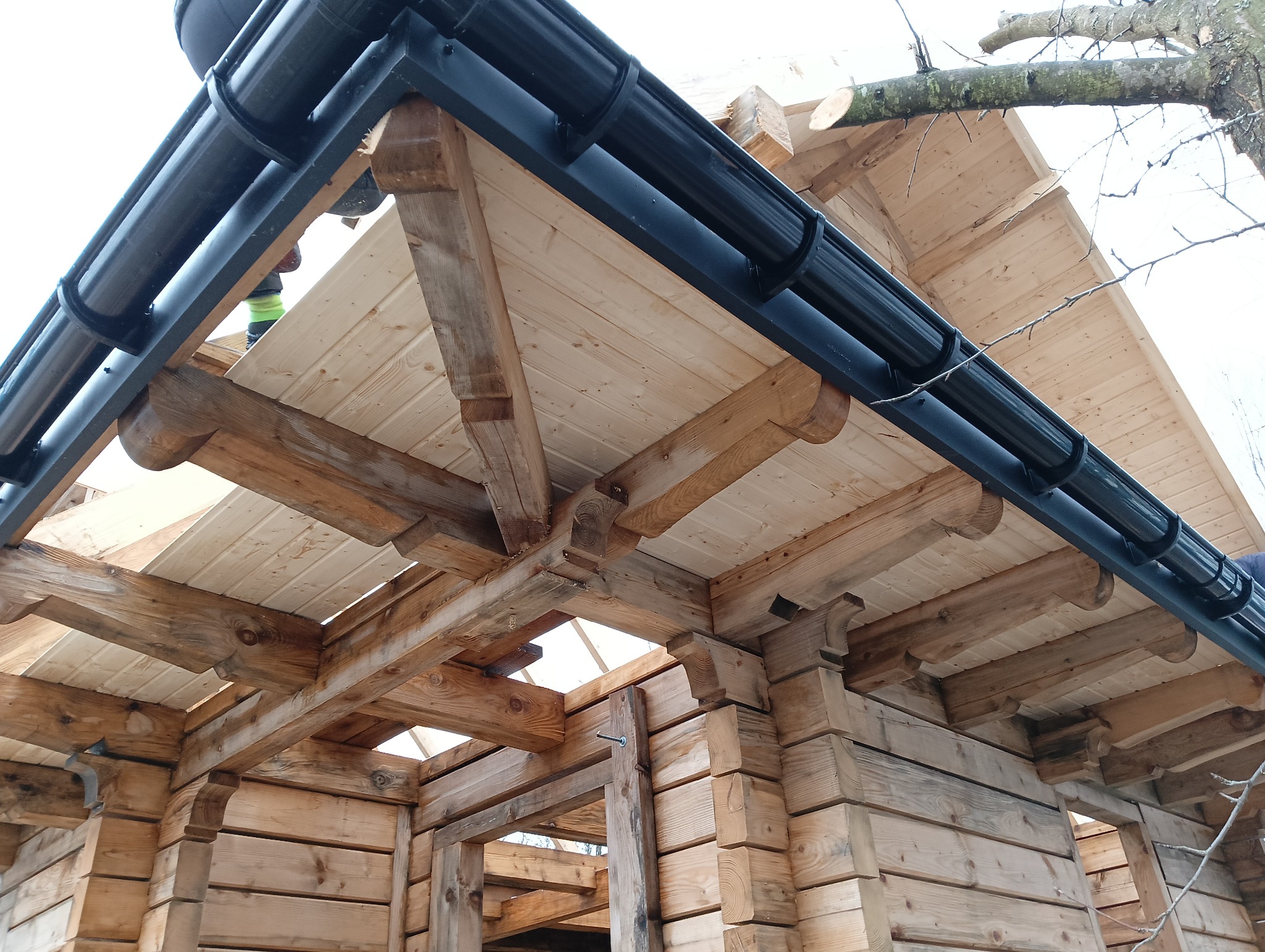 Wnętrze drewnianej konstrukcji dachu z widocznymi elementami więźby i zamontowanymi czarnymi rynnami, fragment drzewa w tle.