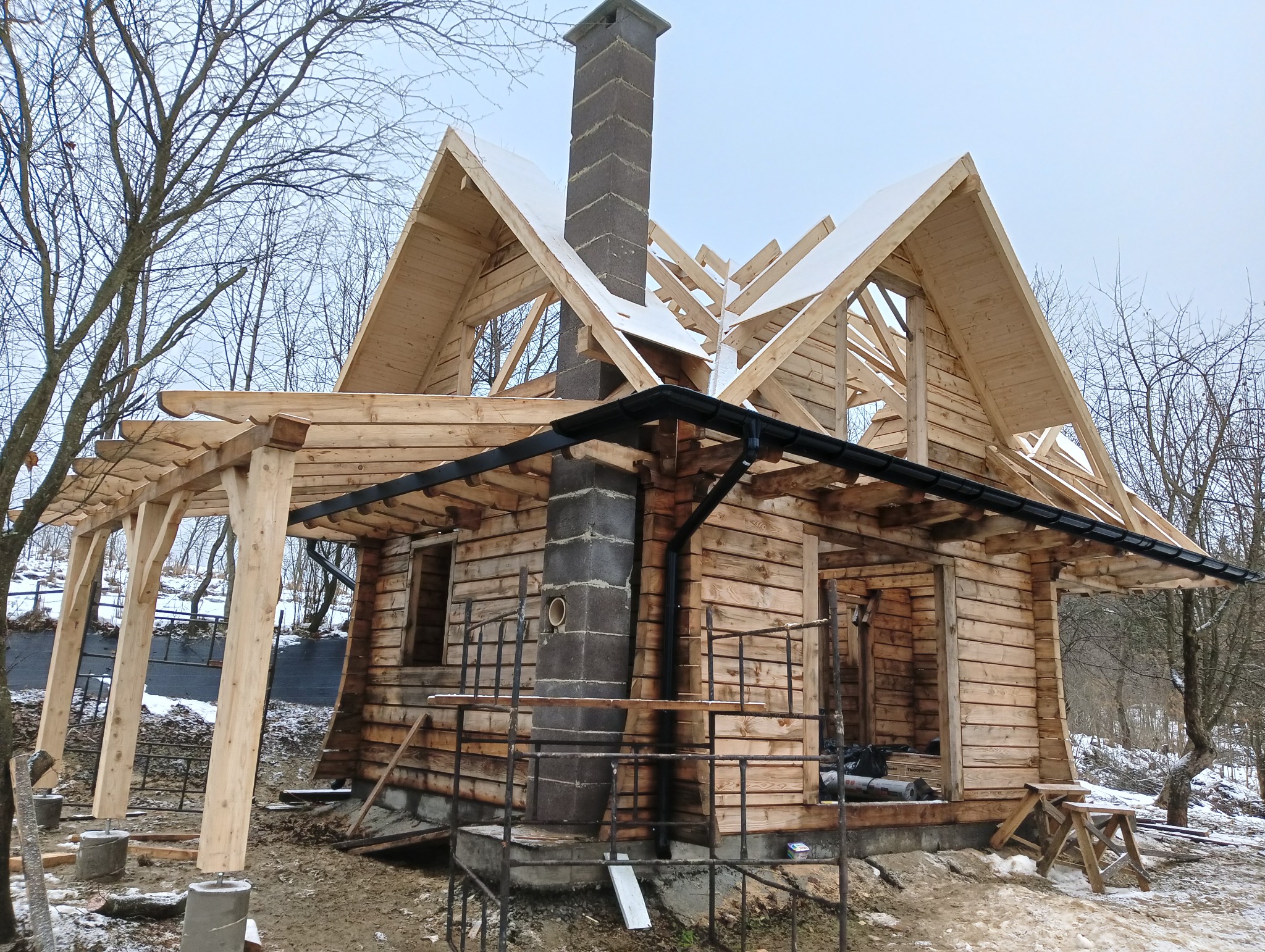 Surowa drewniana konstrukcja domu z widoczną więźbą dachową i kominem, z częściowo zamontowanymi czarnymi rynnami, w zimowej scenerii.