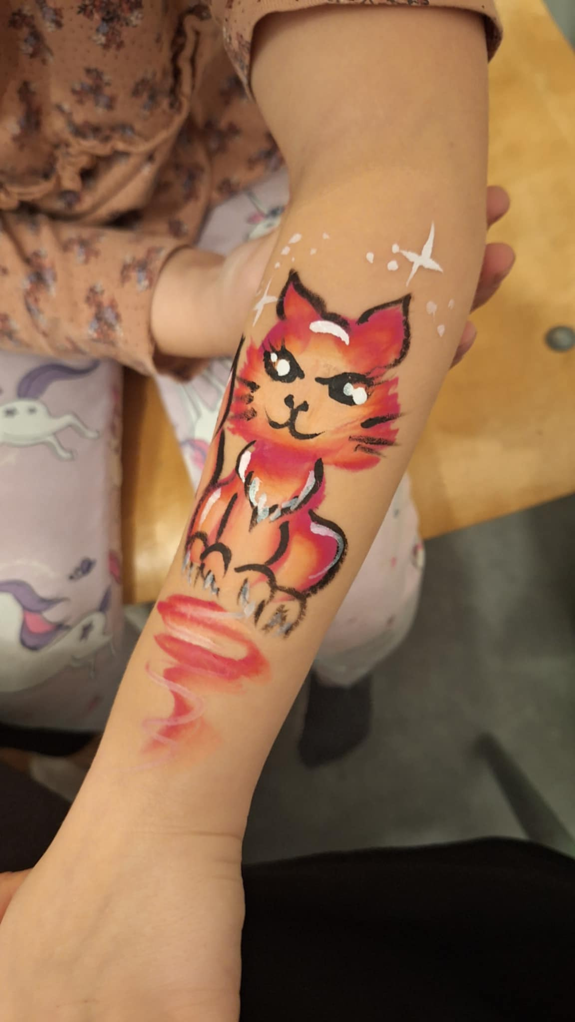 Dziecięca ręka z namalowanym na skórze pomarańczowo-czarnym kotem z białymi akcentami i błyszczącymi gwiazdkami w tle.