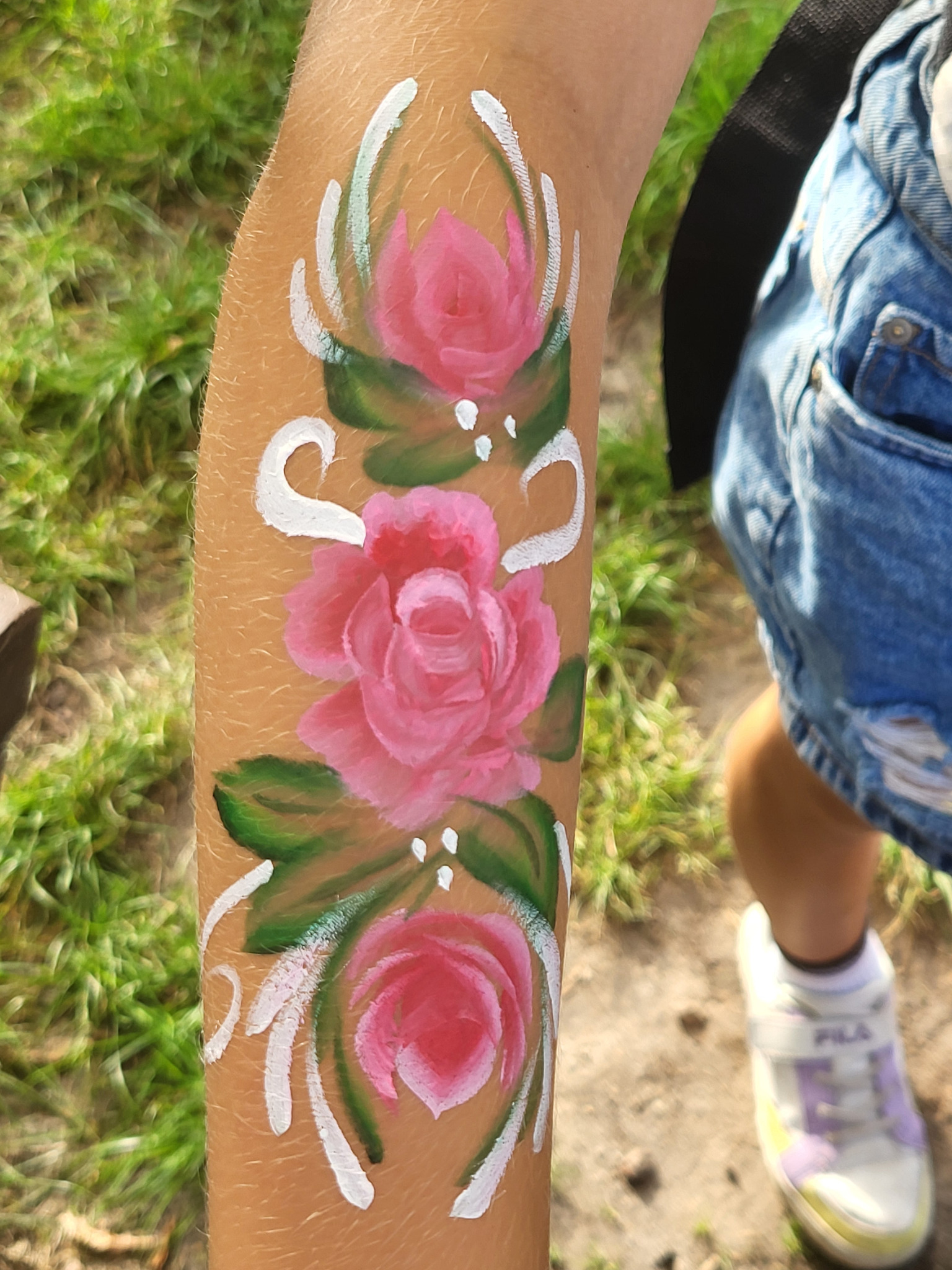 Ręka dziecka z namalowanymi różowymi różami i białymi ornamentami, widoczne fragmenty dżinsowych spodenek i buta sportowego.