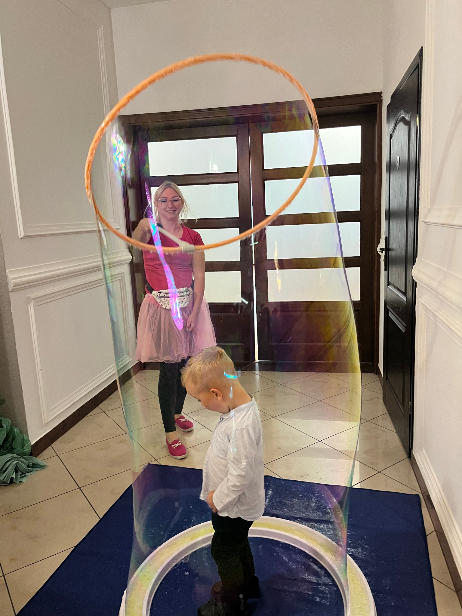 Kobieta w różowej spódniczce tworzy ogromną bańkę mydlaną, w której stoi mały chłopiec, na tle korytarza z ciemnymi drzwiami.