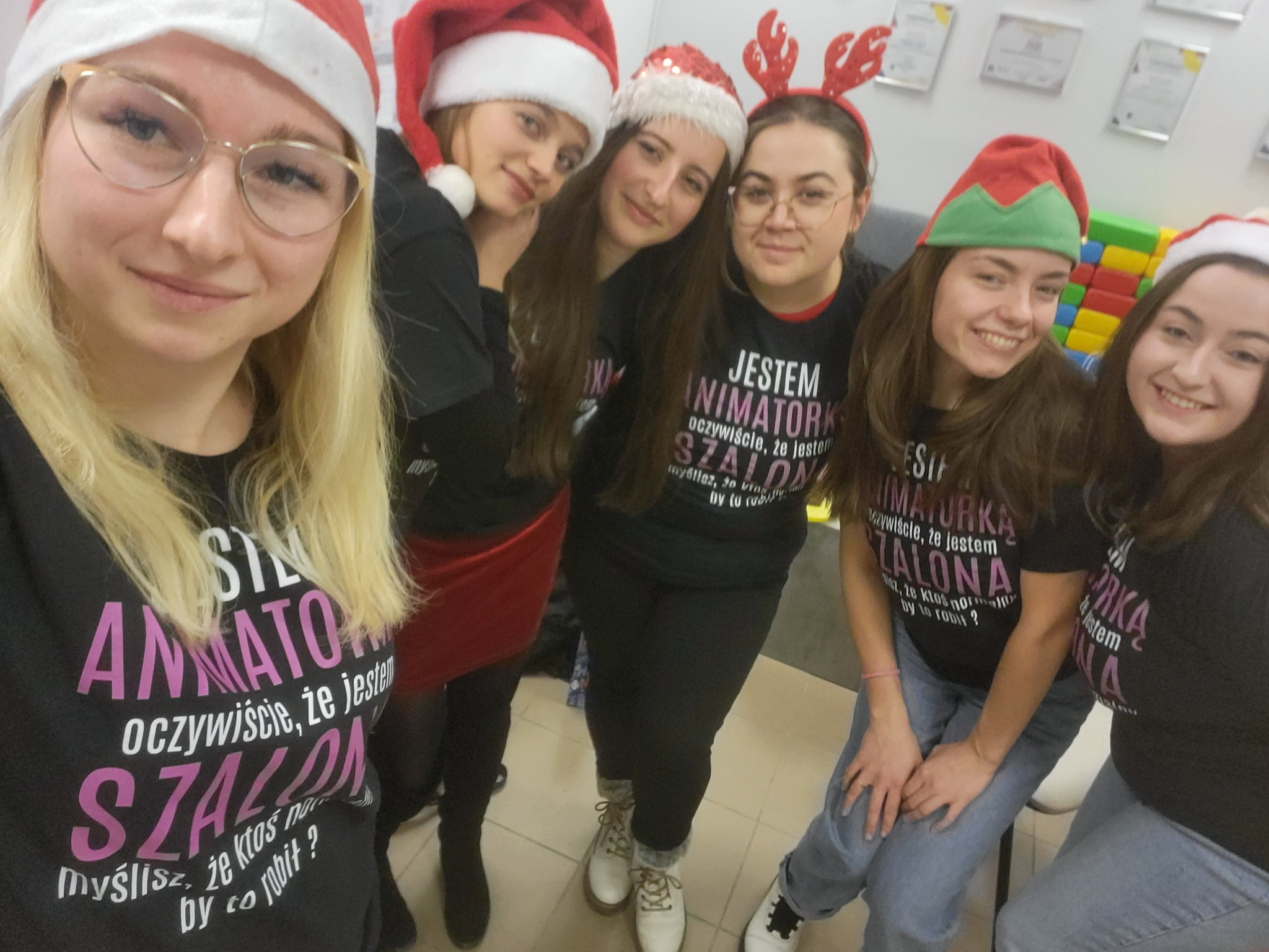 Grupa uśmiechniętych animatorek w świątecznych czapkach i koszulkach z napisem 'Jestem animatorką, oczywiście, że jestem szalona', pozuje do selfie na tle ściany z dyplomami i kolorowymi klockami.