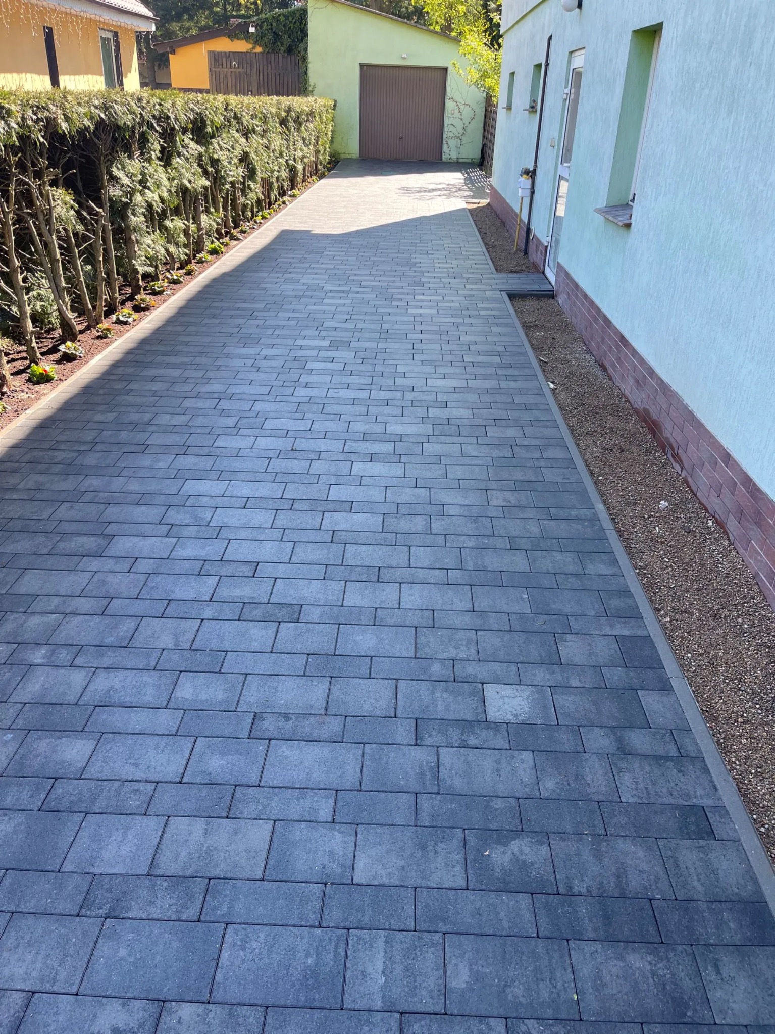Długa, prosta ścieżka z szarej kostki brukowej prowadząca do garażu, obramowana żywopłotem i ścianą budynku.