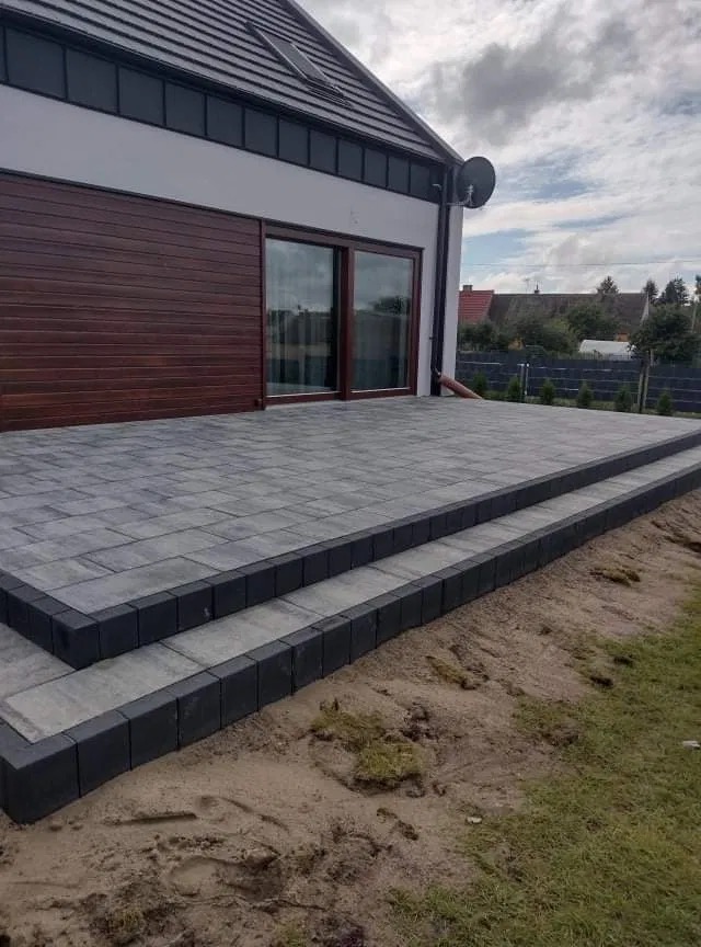 Szara kostka brukowa ułożona na tarasie przed domem z dwoma stopniami, obrzeża wykończone ciemniejszą kostką.