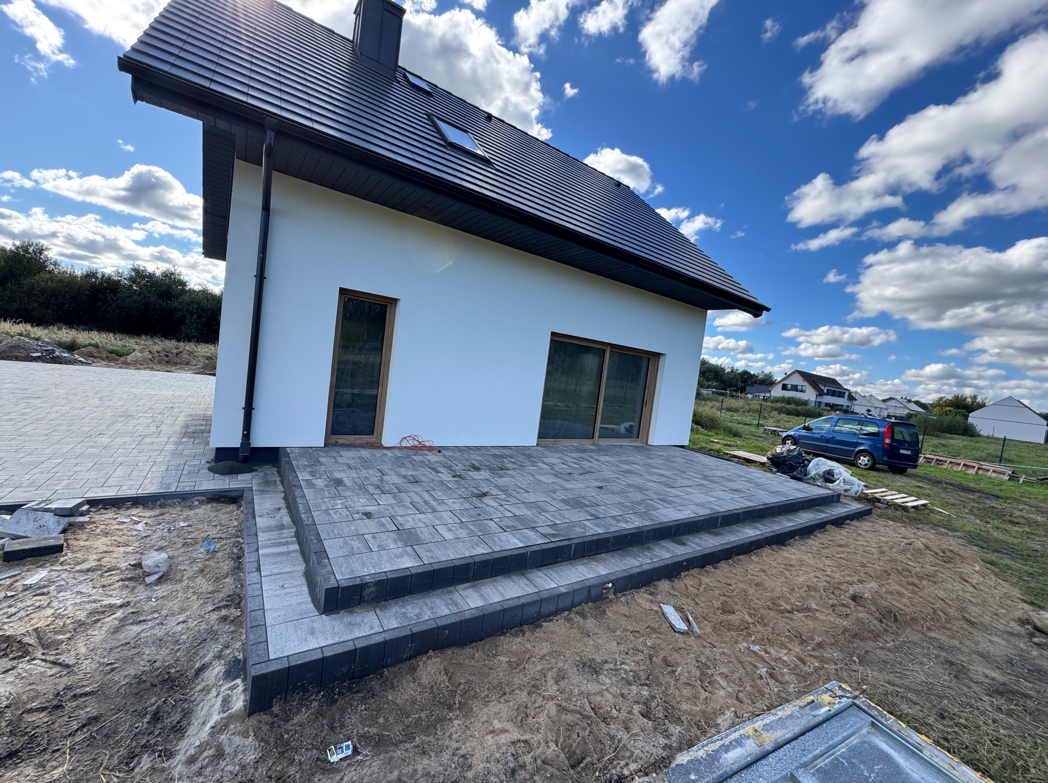 Nowoczesny taras z kostki brukowej w odcieniach szarości, z dwoma stopniami, przylegający do białego domu z ciemną dachówką. Widok na częściowo zagospodarowany teren.