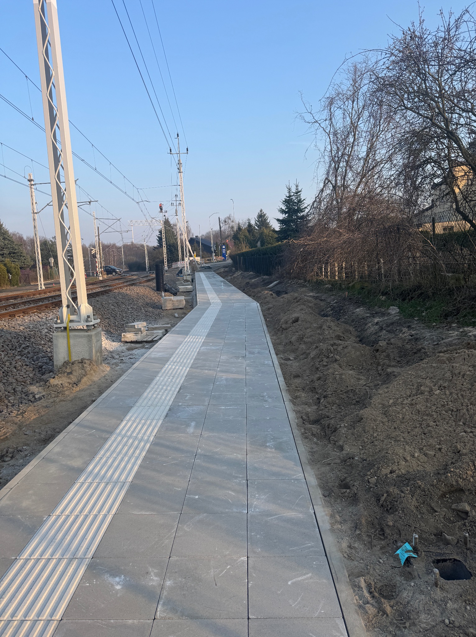 Nowa, szara nawierzchnia z kostki brukowej z pasem dla niewidomych, ułożona wzdłuż torów kolejowych. Widoczne słupy trakcyjne i świeżo obsypana ziemia.