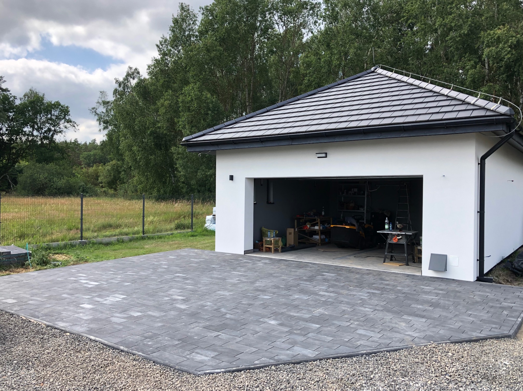 Nowoczesny podjazd z szarej kostki brukowej ułożonej wzorem jodełki, obok garażu z białymi ścianami i ciemną dachówką. Otoczenie: trawa, drzewa, żwir.