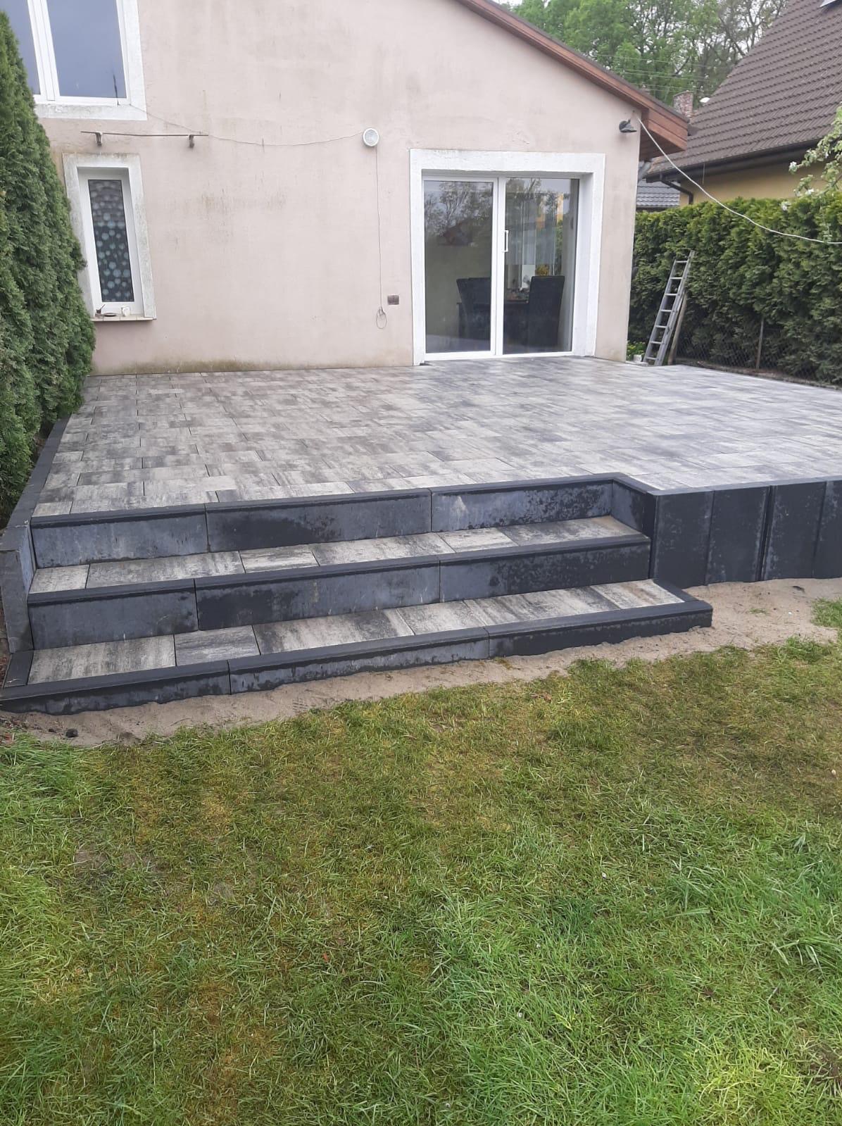 Wykończony taras z kostki brukowej z trzema schodami, przylegający do budynku z oknami i drzwiami przesuwnymi. Widoczny trawnik i drabina oparta o żywopłot.