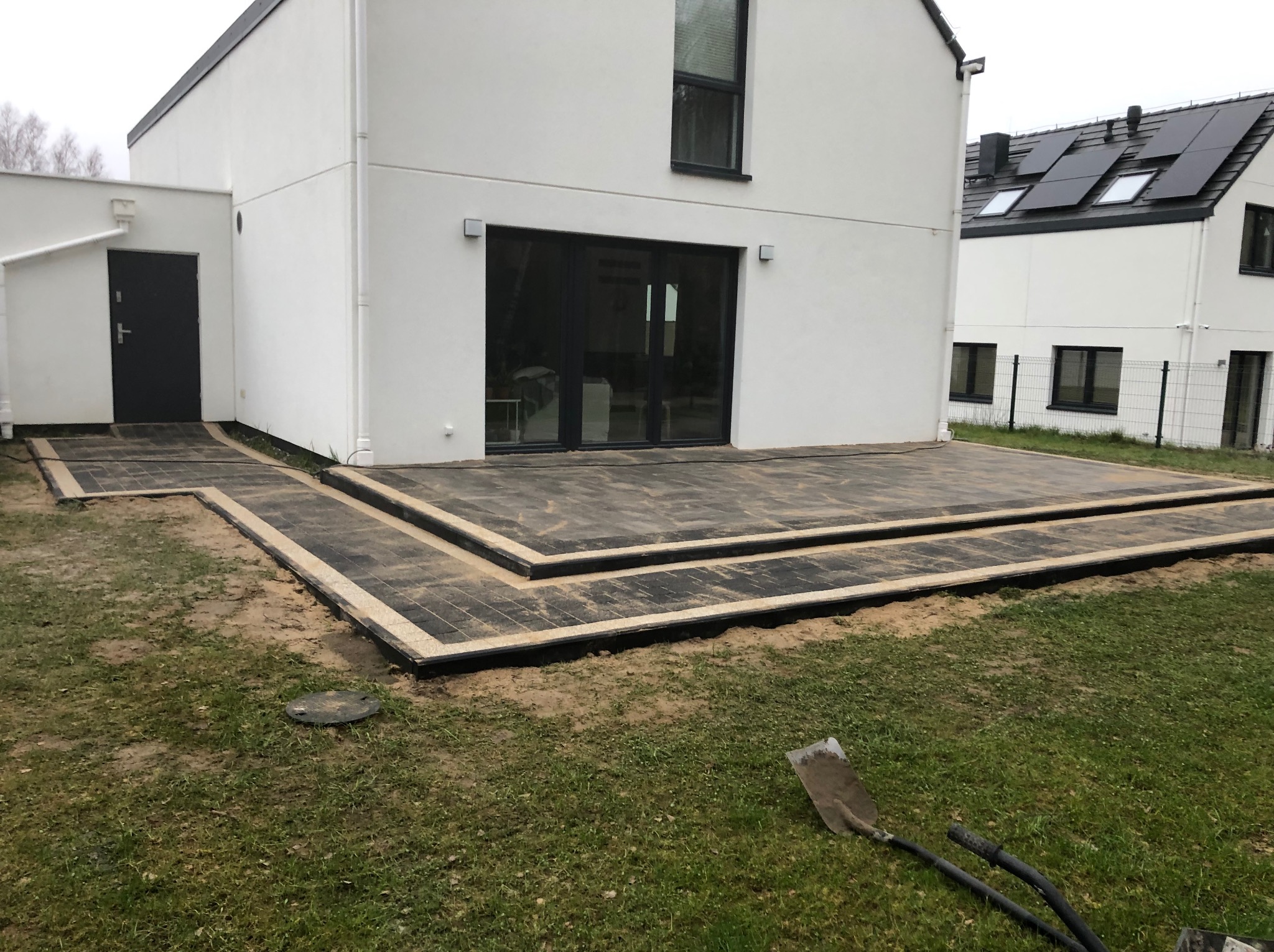 Ułożona kostka brukowa tworząca taras przy nowoczesnym, białym domu z czarnymi oknami i drzwiami. Widoczna trawa i narzędzia brukarskie.