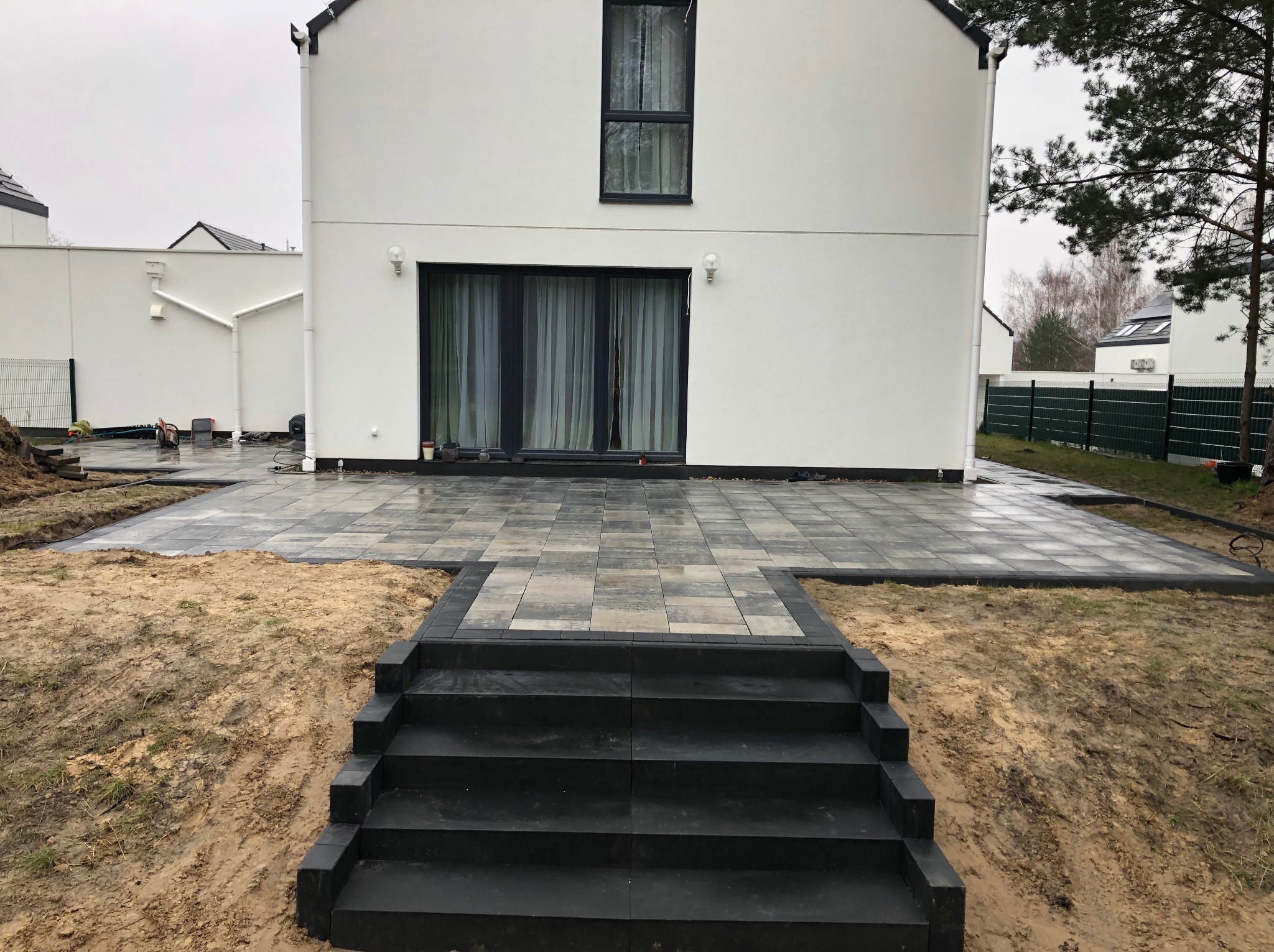 Nowoczesny taras z szarej kostki brukowej prowadzący do białego domu z dużymi oknami, schody z kostki, surowy krajobraz.