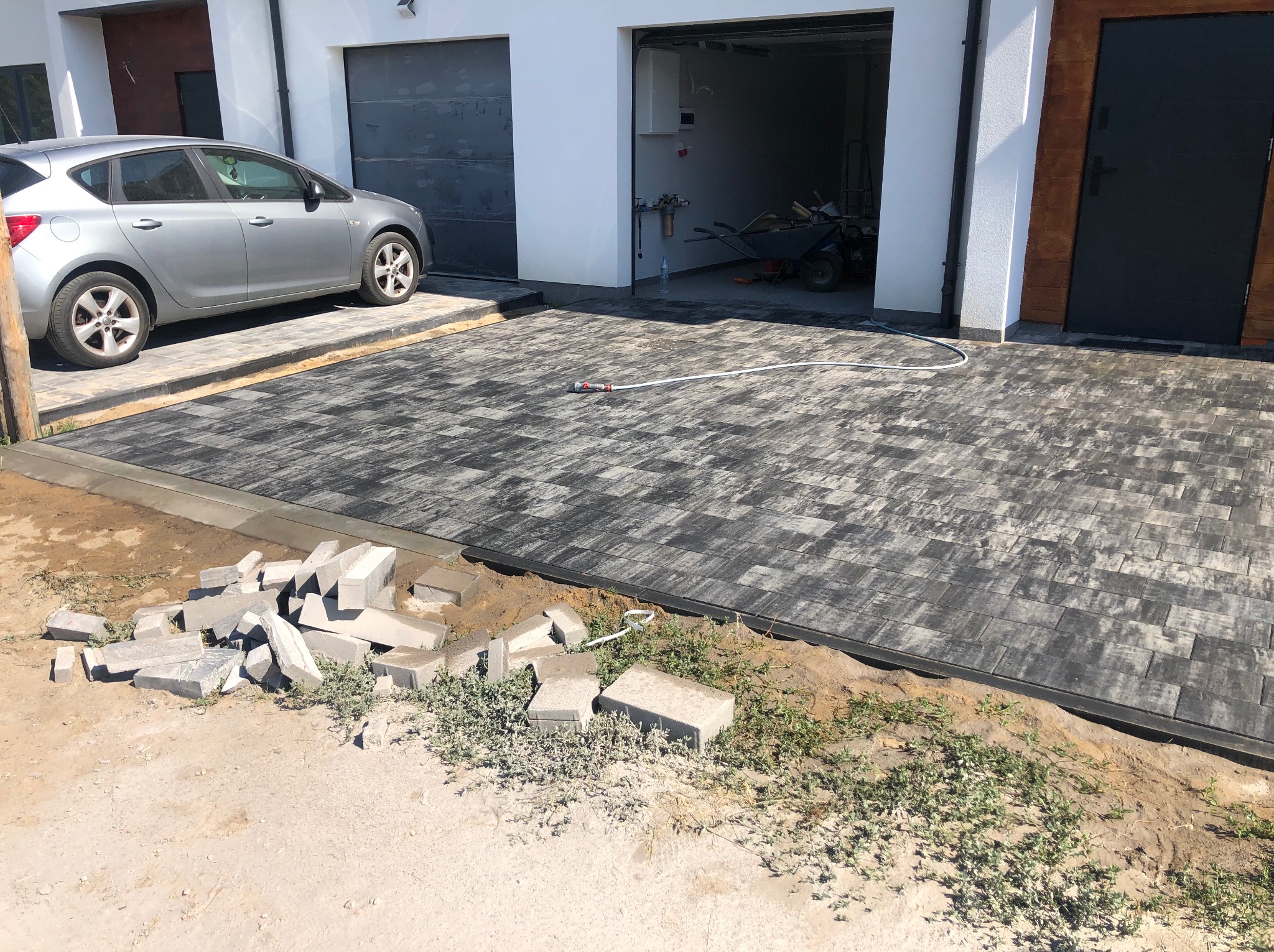 Częściowo ułożona, grafitowa kostka brukowa przed garażem i domem jednorodzinnym, z widocznym stosem kostki do ułożenia. W tle zaparkowany samochód osobowy.