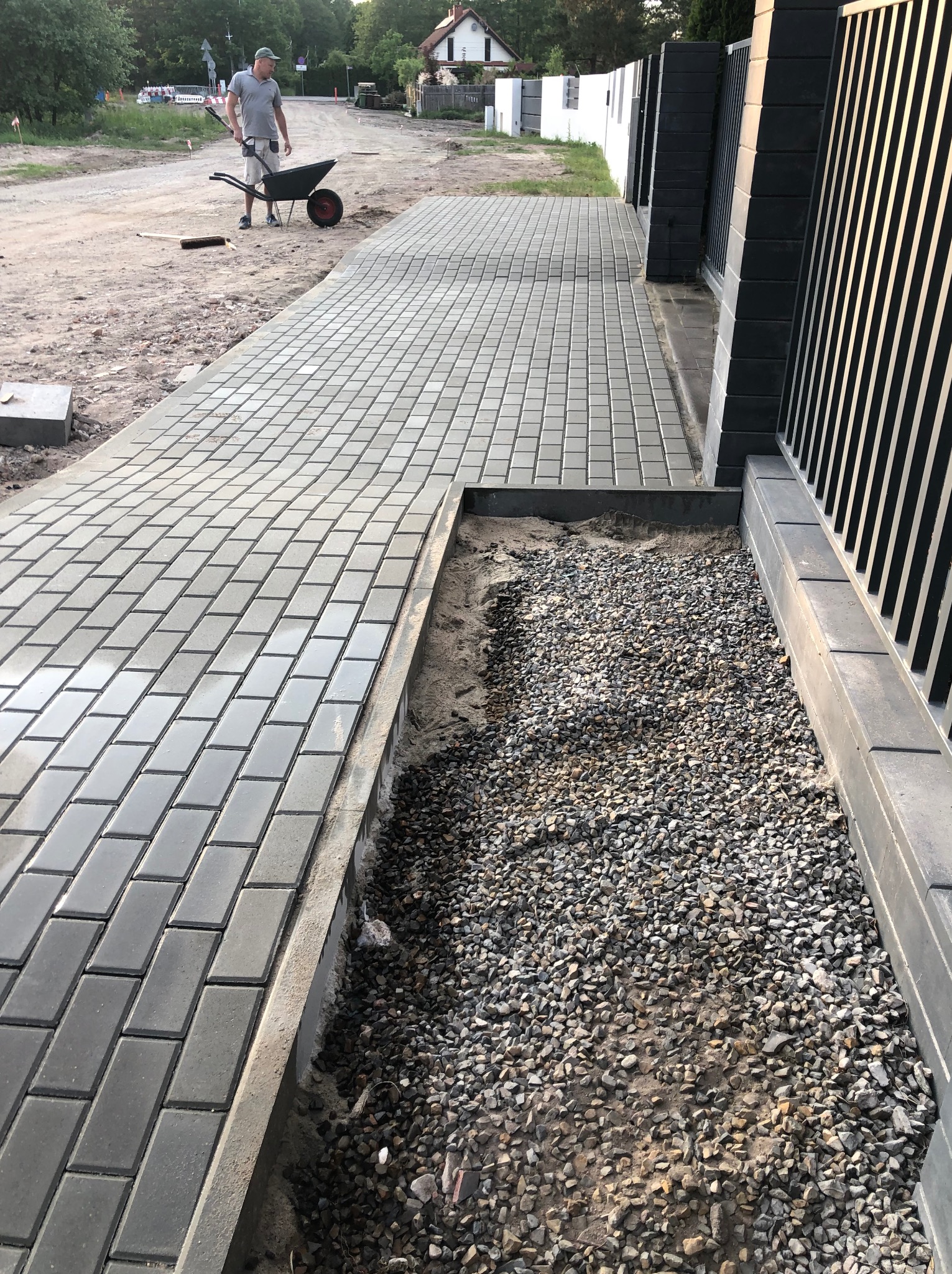 Świeżo ułożona, szara kostka brukowa tworząca ścieżkę wzdłuż ogrodzenia. Widoczny fragment przygotowanego podłoża z kamieniami i pracownik z taczką w tle.