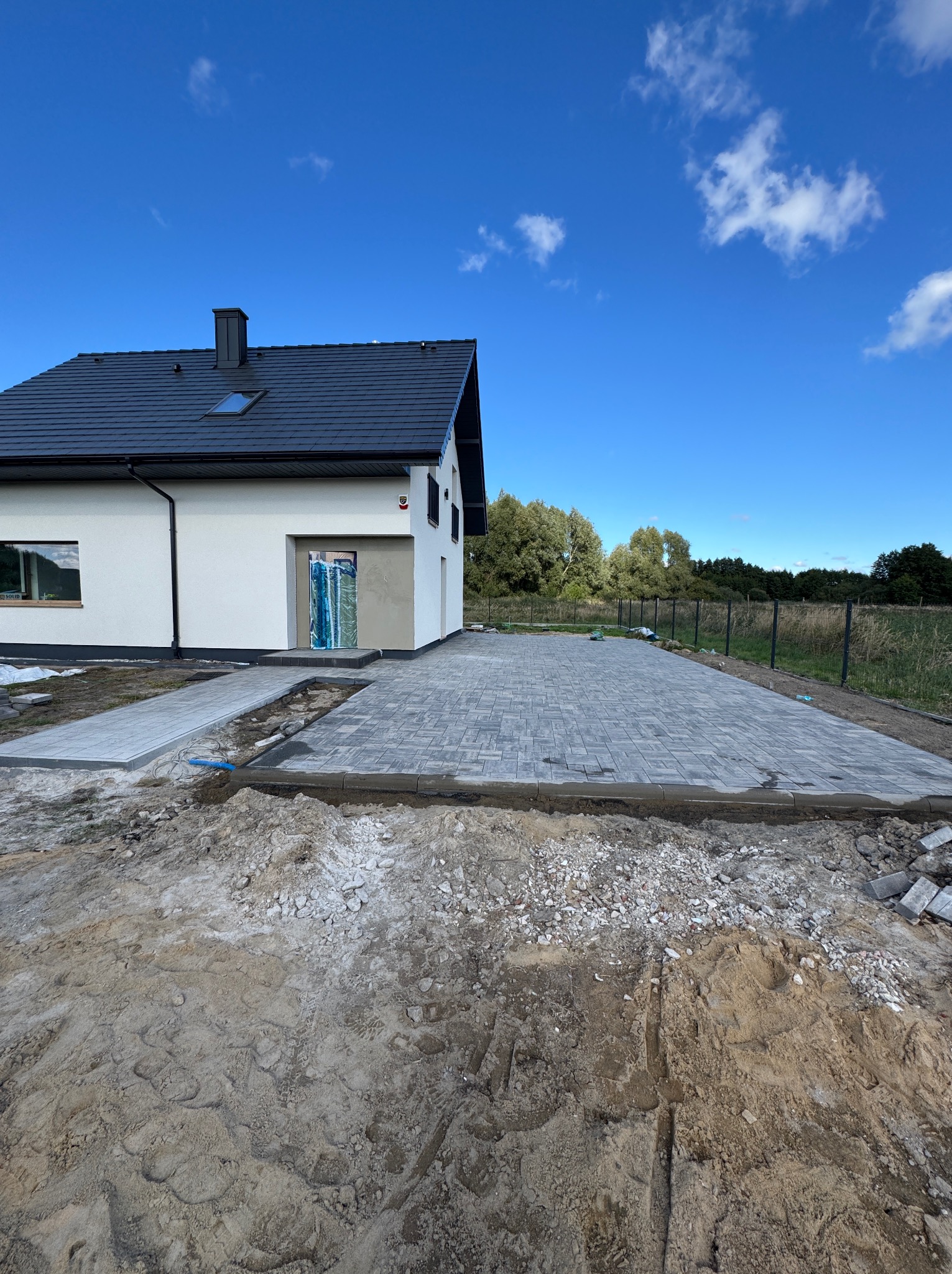 Układanie szarej kostki brukowej na podjeździe przed nowoczesnym domem z czarnym dachem i niebieskim niebem w tle. Widoczny etap prac, przygotowane podłoże.