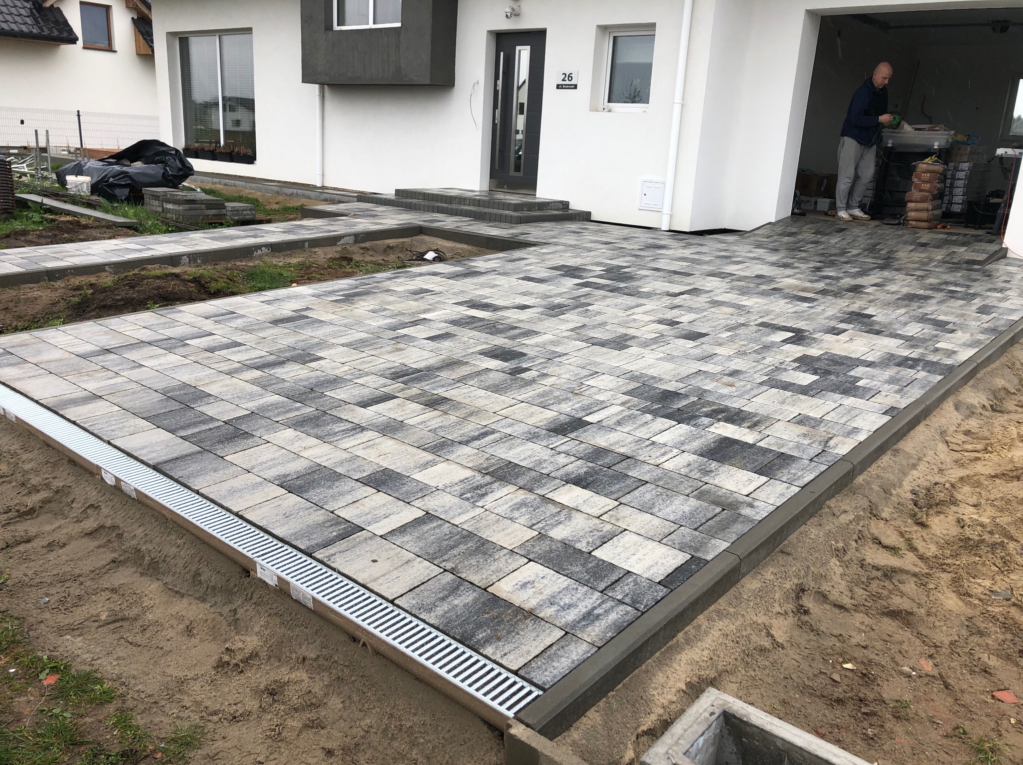 Ułożona kostka brukowa przed domem z numerem 26, widoczne odwodnienie liniowe i fragment brukarza w garażu. Nowoczesna aranżacja przestrzeni.