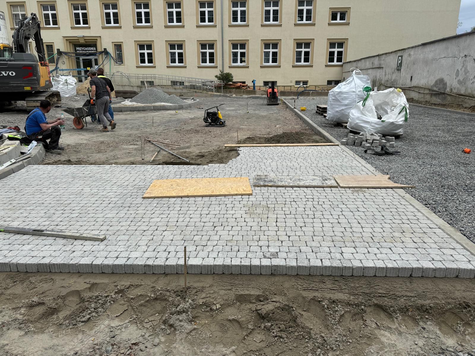 Układanie szarej kostki brukowej na terenie zewnętrznym, widoczne narzędzia, materiały i ekipa pracująca; w tle budynek przychodni.