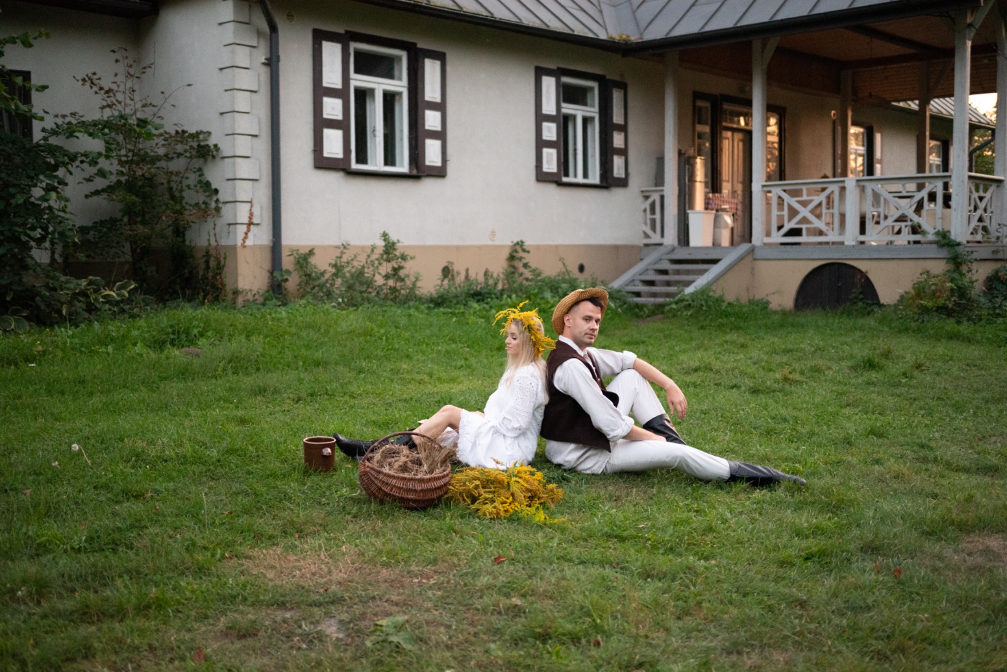 Para w strojach ludowych siedzi plecami do siebie na trawie przed wiejskim domem. Koszyk, wiązanka kwiatów i kubek obok nich.