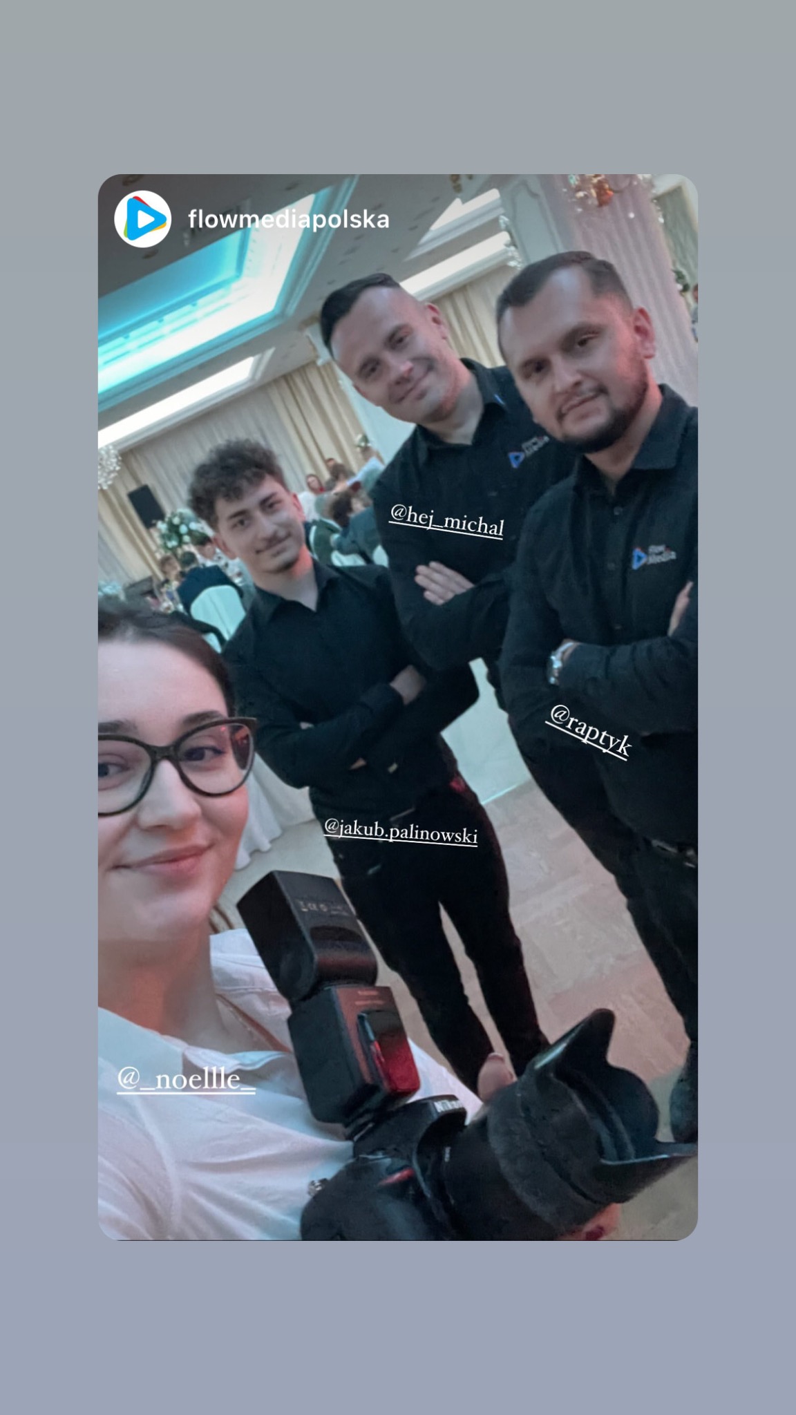 Grupa fotografów w ciemnych koszulach z logo firmy, pozujących do selfie z aparatem i lampą błyskową na tle sali bankietowej.