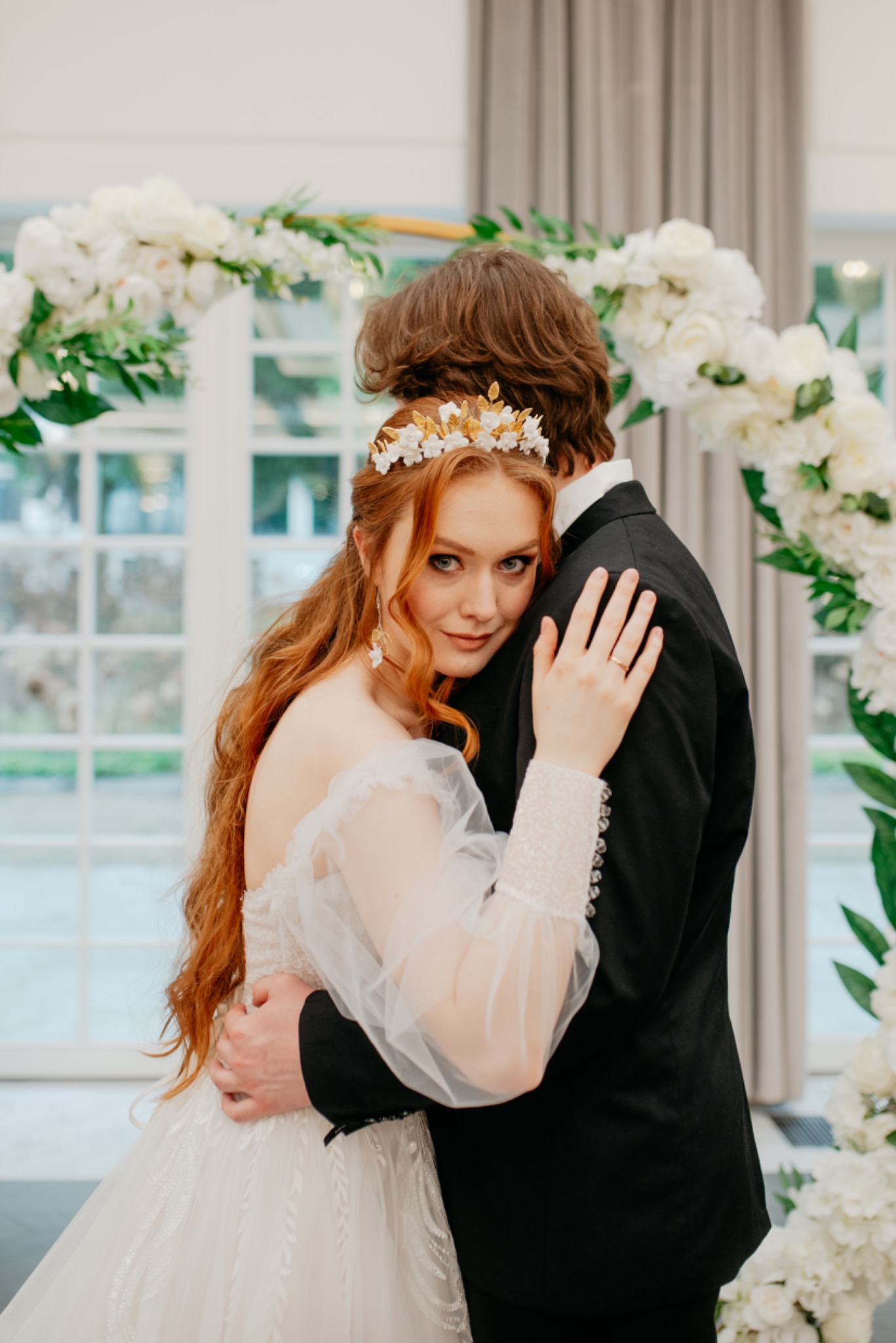 Panna młoda z rudymi włosami i złotą koroną obejmuje pana młodego na tle białego łuku kwiatowego w jasnym wnętrzu.