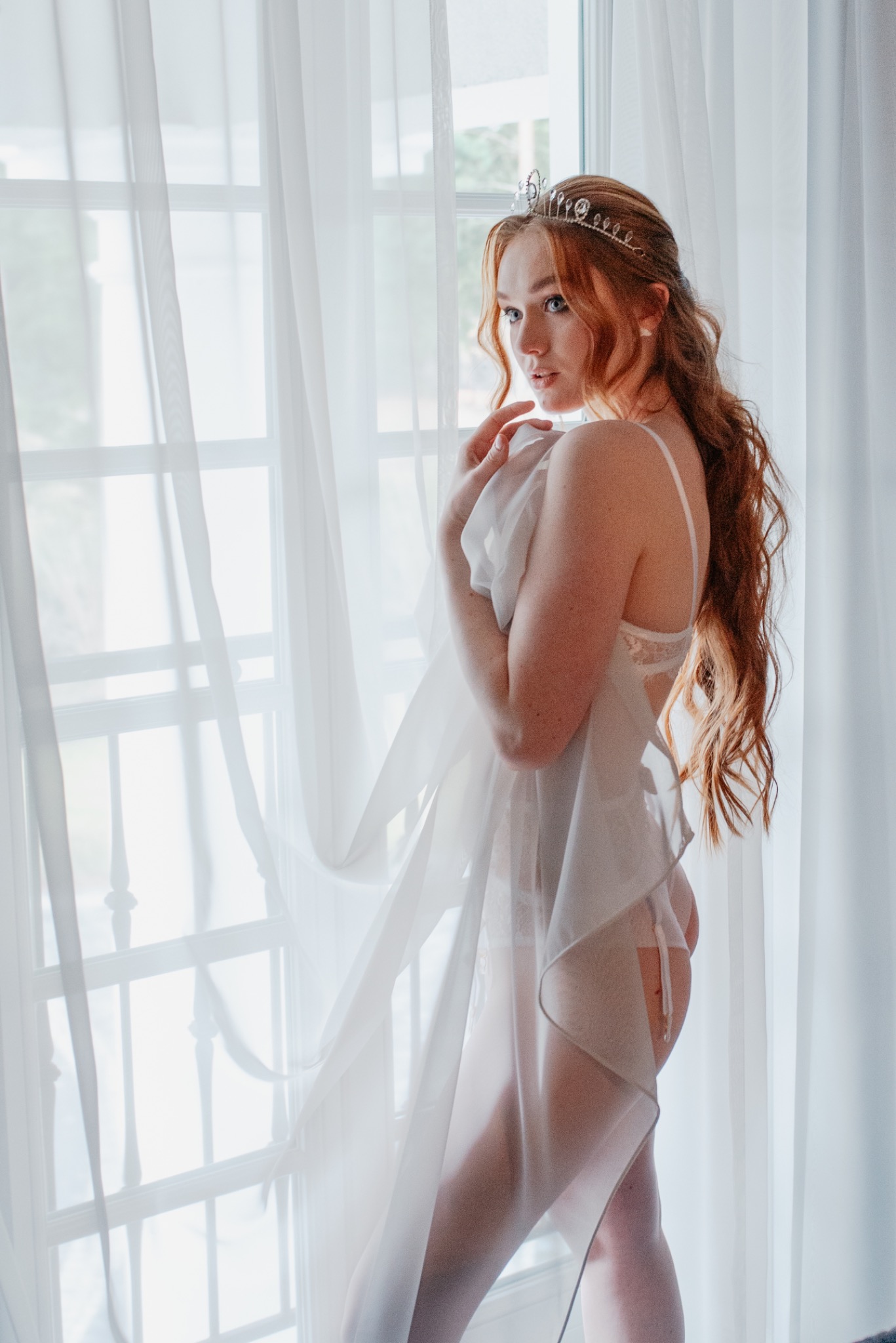 Ginger woman with tiara posing in lingerie and sheer fabric by a window with white curtains.