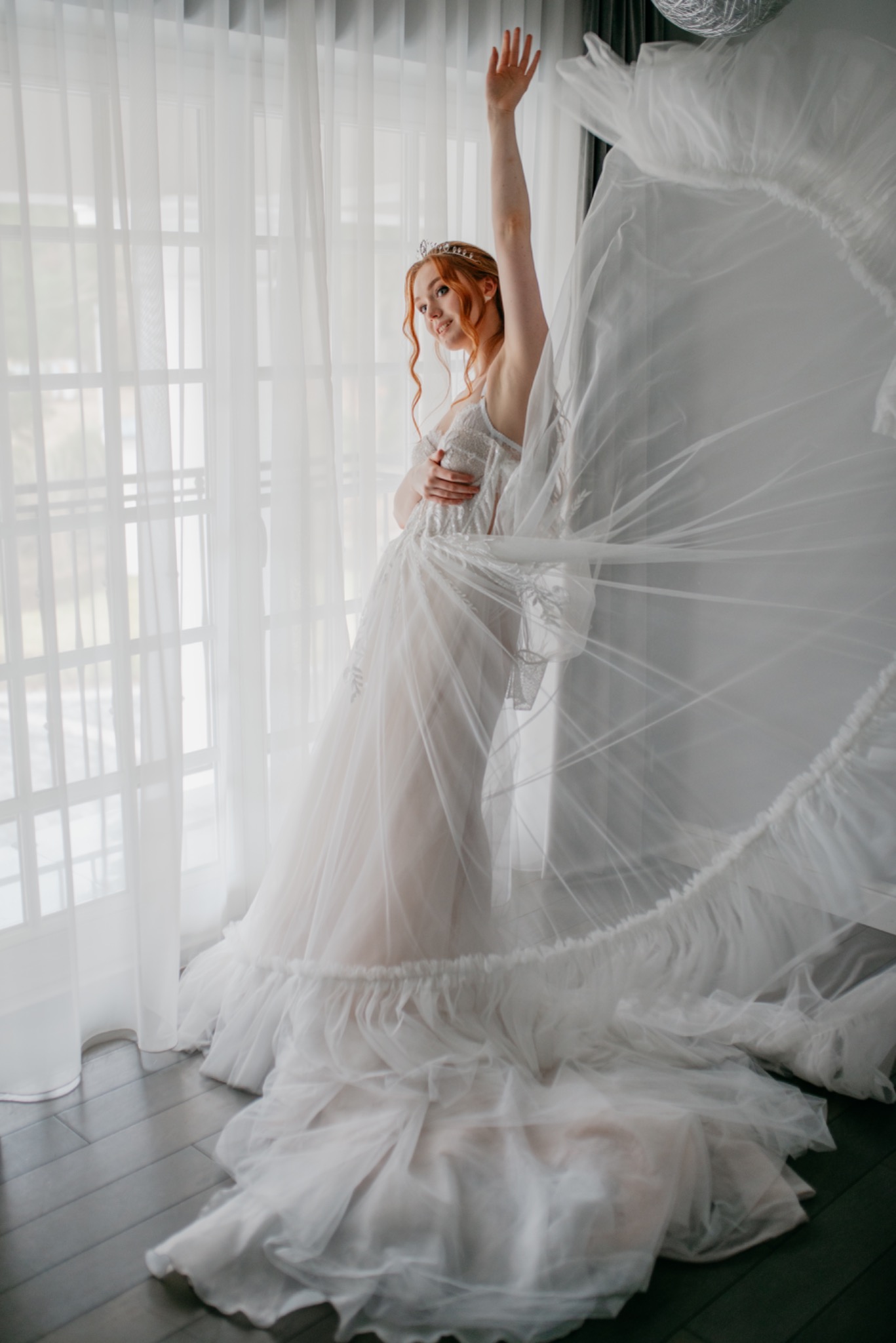 Uśmiechnięta panna młoda z rudymi włosami i tiarą, pozuje w zwiewnej sukni ślubnej przy oknie, z uniesioną ręką i fragmentem falującego materiału sukni.