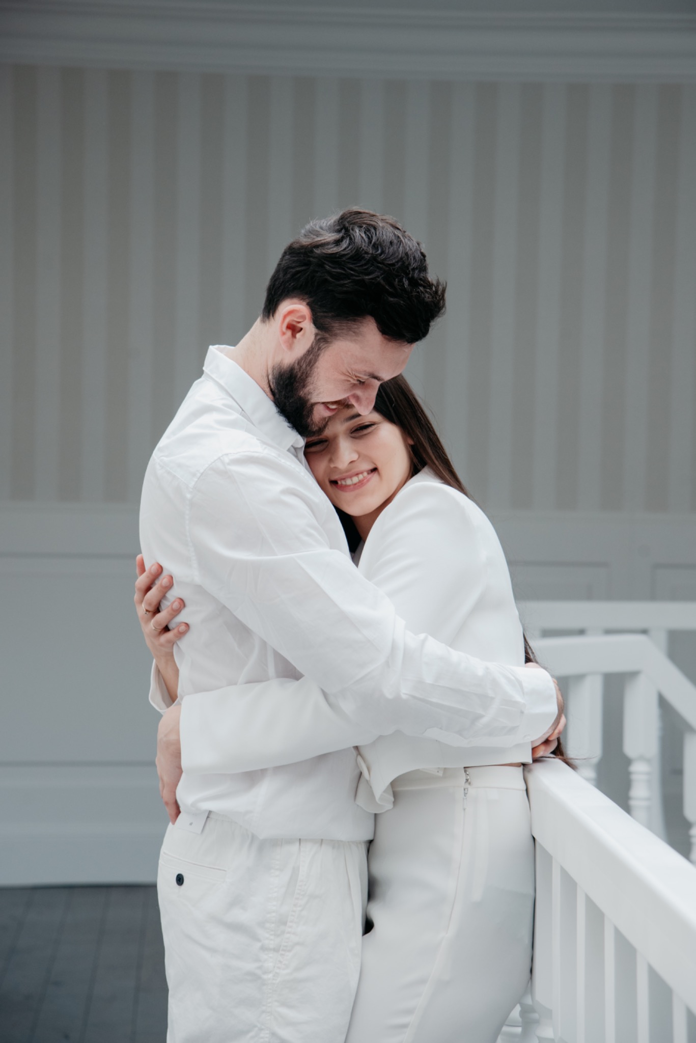 Uśmiechnięta para w białych ubraniach, obejmująca się na tle jasnego, minimalistycznego wnętrza z białą balustradą.