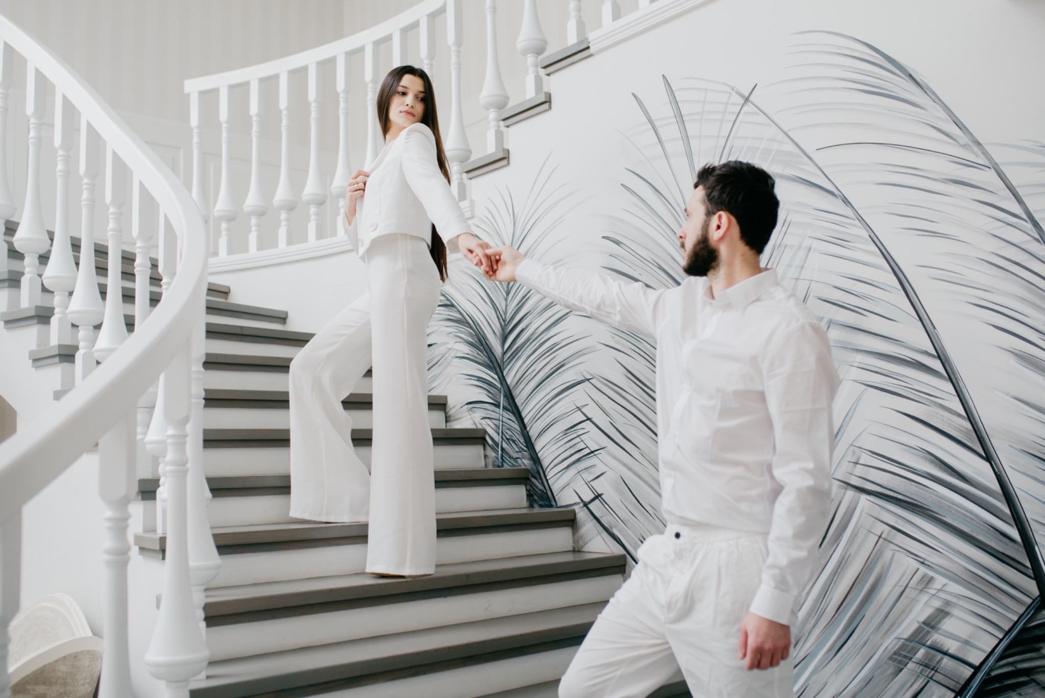 Para w bieli na schodach z dekoracyjną tapetą w tle, kobieta spogląda przez ramię, mężczyzna trzyma ją za rękę.