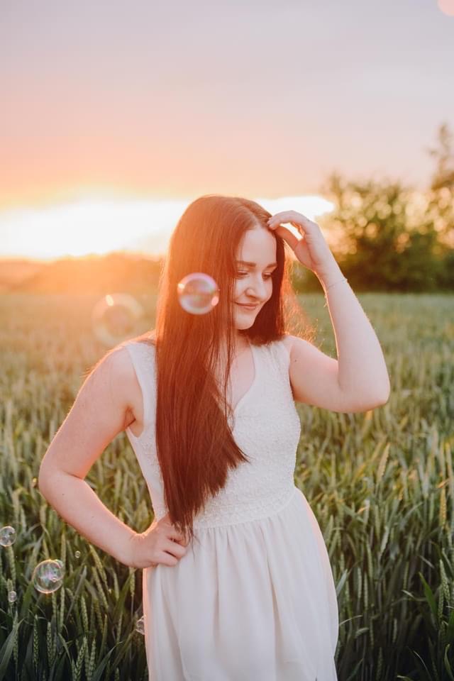 Młoda kobieta w białej sukience pozuje na tle pola pszenicy o zachodzie słońca, z bańkami mydlanymi unoszącymi się w powietrzu.