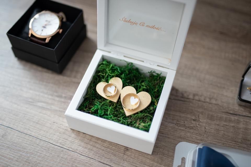Złote obrączki ślubne w białym, otwartym pudełku z napisem 'Ślubuję Ci miłość...', umieszczone na zielonym mchu i drewnianych sercach, obok czarne pudełko z zegarkiem.