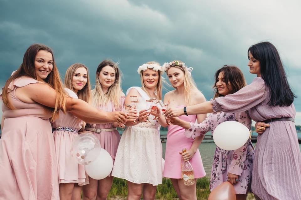 Grupa siedmiu kobiet w pastelowych sukienkach wznosi toast kieliszkami, tło stanowi pochmurne niebo i woda, jedna z kobiet trzyma butelkę różowego wina, a w kadrze widoczne są białe balony.