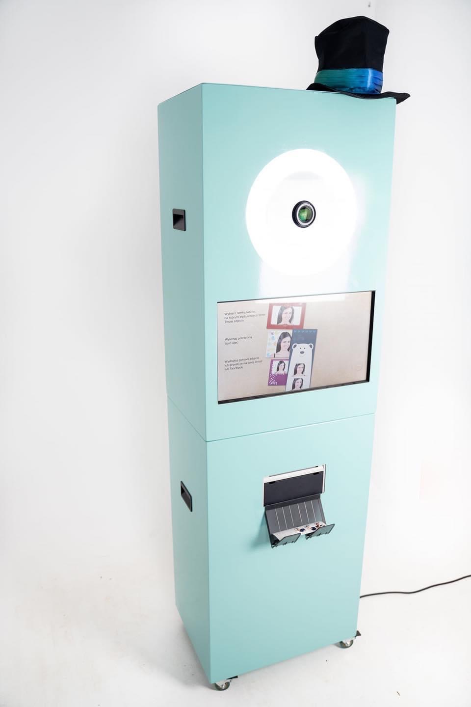 Automat do robienia zdjęć w kolorze miętowym z czarnym cylindrem na górze, ekran dotykowy z wyborem szablonów zdjęć, wysuwana szuflada z wydrukowanymi fotografiami.