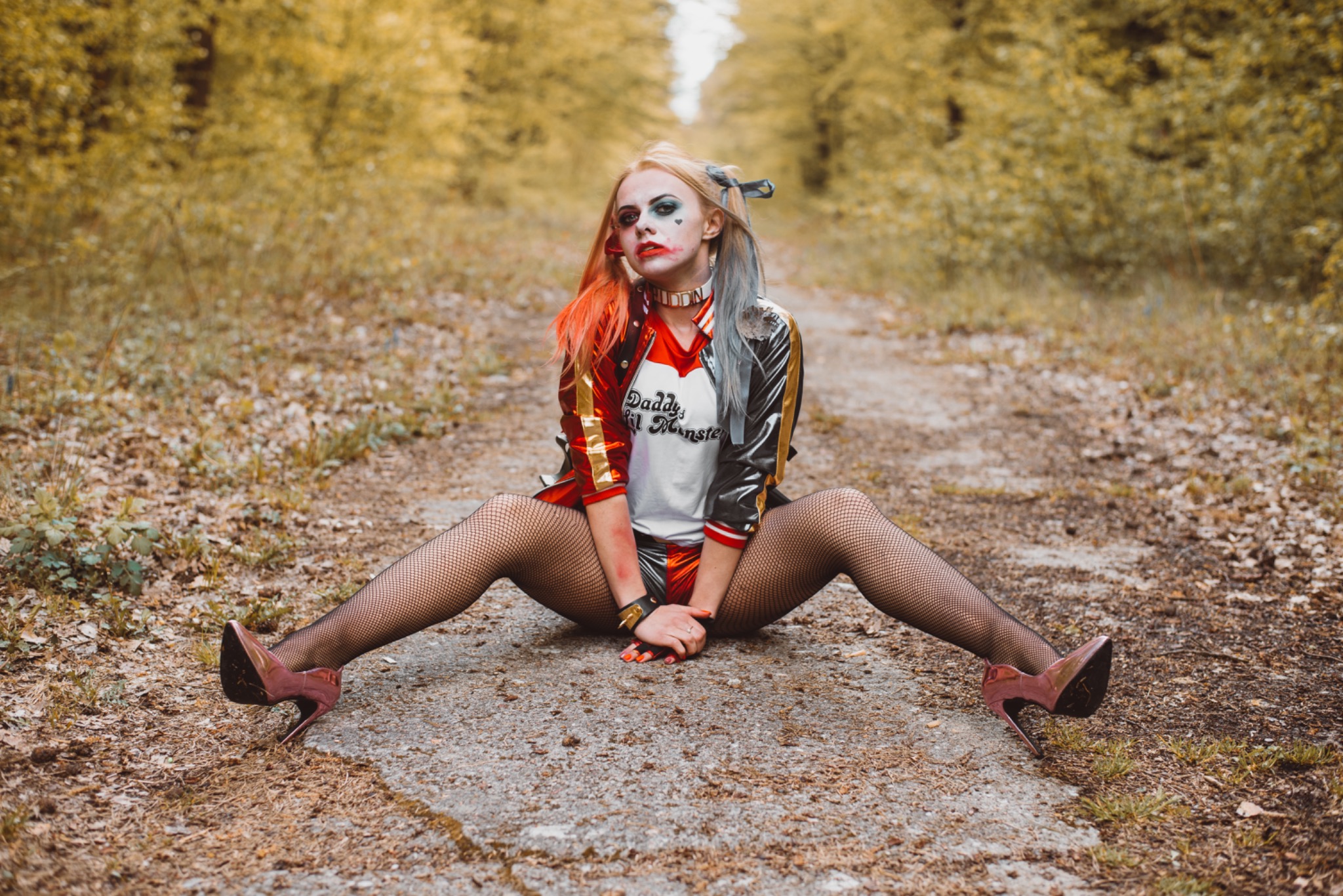 Kobieta w kostiumie Harley Quinn siedzi na leśnej ścieżce, pozuje z rozłożonymi nogami, widać rajstopy kabaretki i lakierowane szpilki, tło stanowią drzewa w jesiennych kolorach.