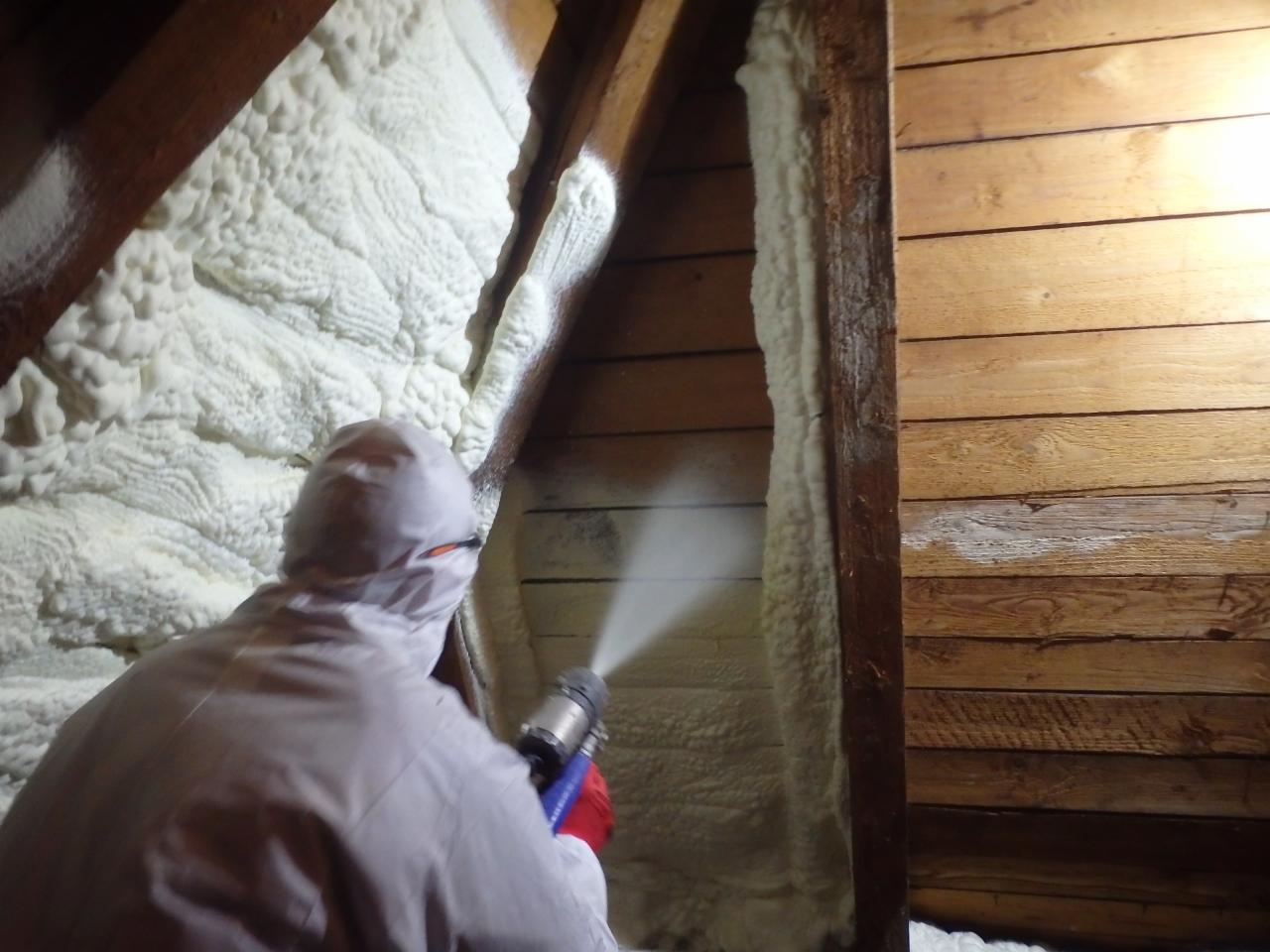 Pracownik w kombinezonie ochronnym aplikuje izolację natryskową na poddaszu, widoczna struktura piany i drewniana konstrukcja dachu.
