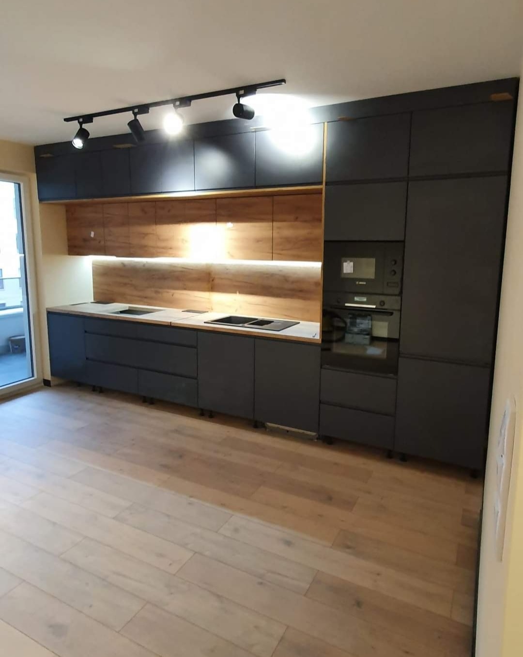 Nowoczesna kuchnia z czarnymi szafkami i drewnianym panelem nad blatem, oświetlona reflektorami sufitowymi i listwą LED, z widocznymi elementami zabudowy AGD, na jasnej drewnianej podłodze.