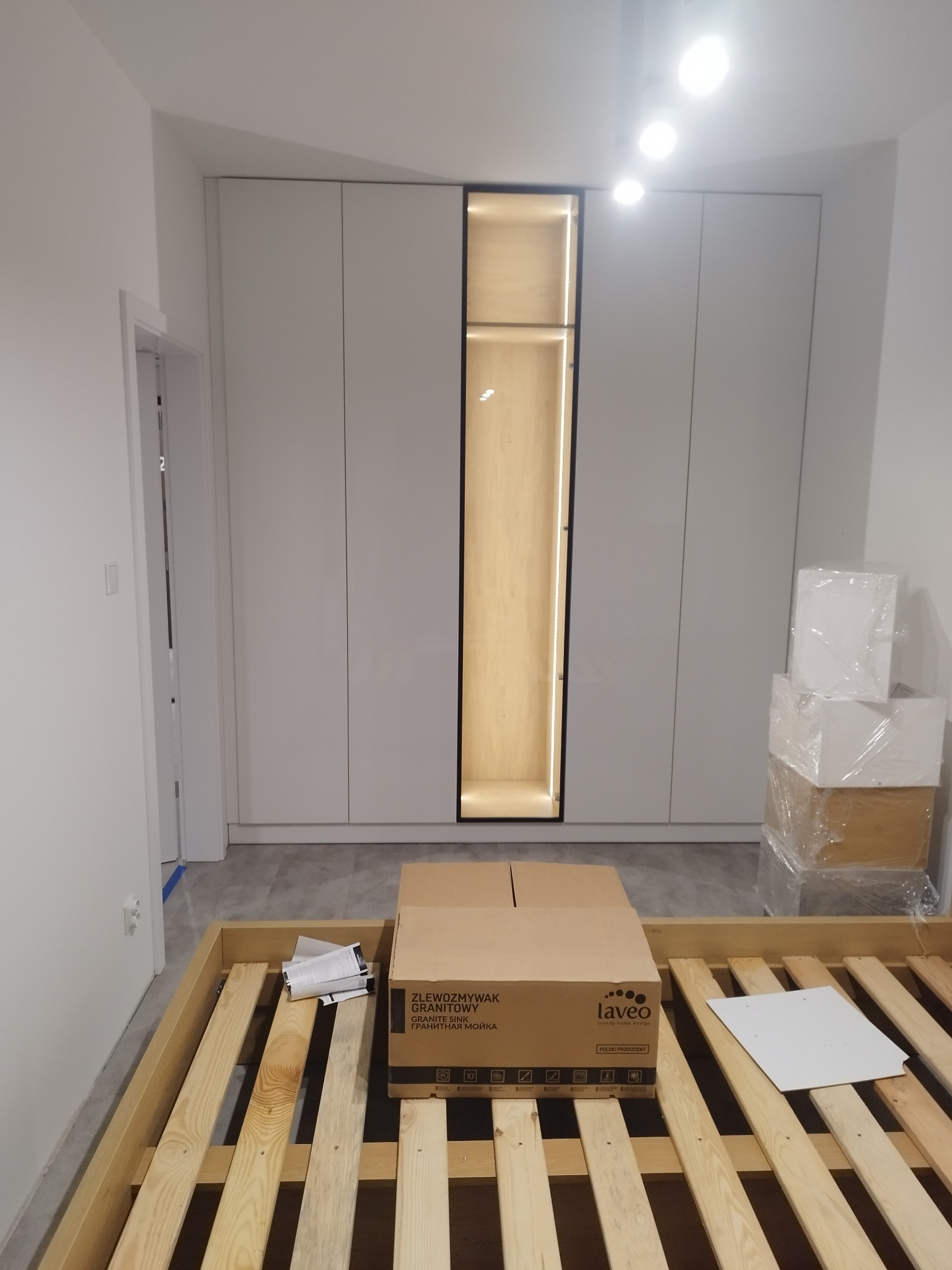 Biała szafa wnękowa z podświetlaną wnęką, karton ze zlewozmywakiem i stelaż łóżka w surowym wnętrzu.