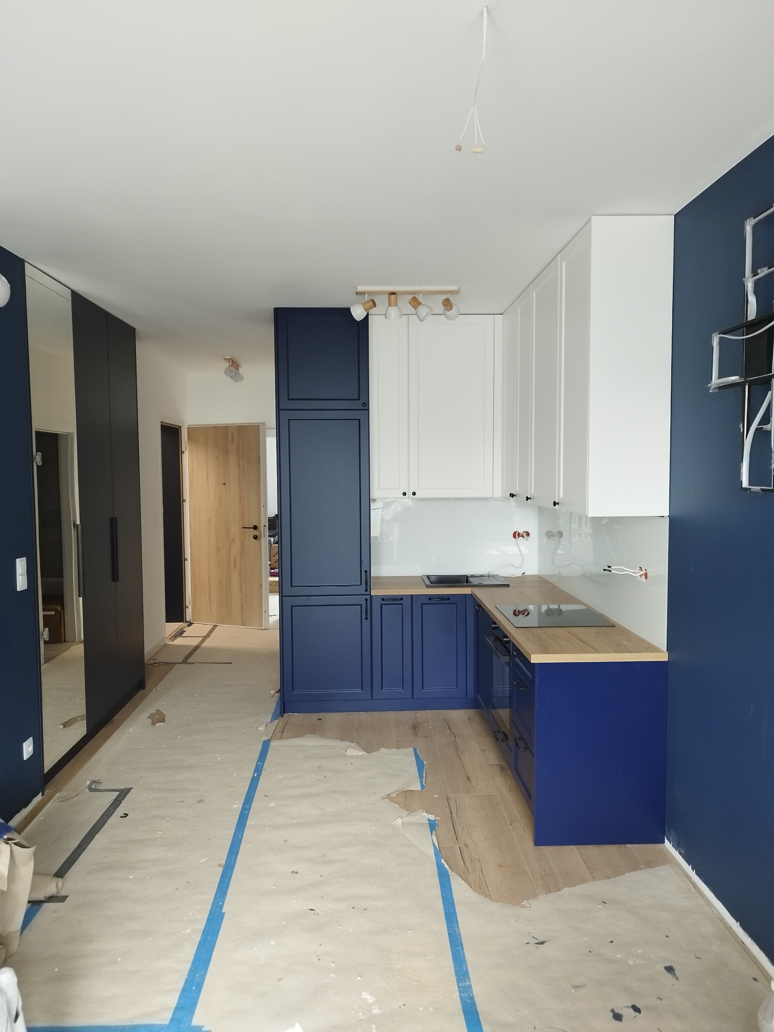 Niebiesko-biała kuchnia w trakcie montażu, zabezpieczona podłoga, widoczne niedokończone elementy oświetlenia.
