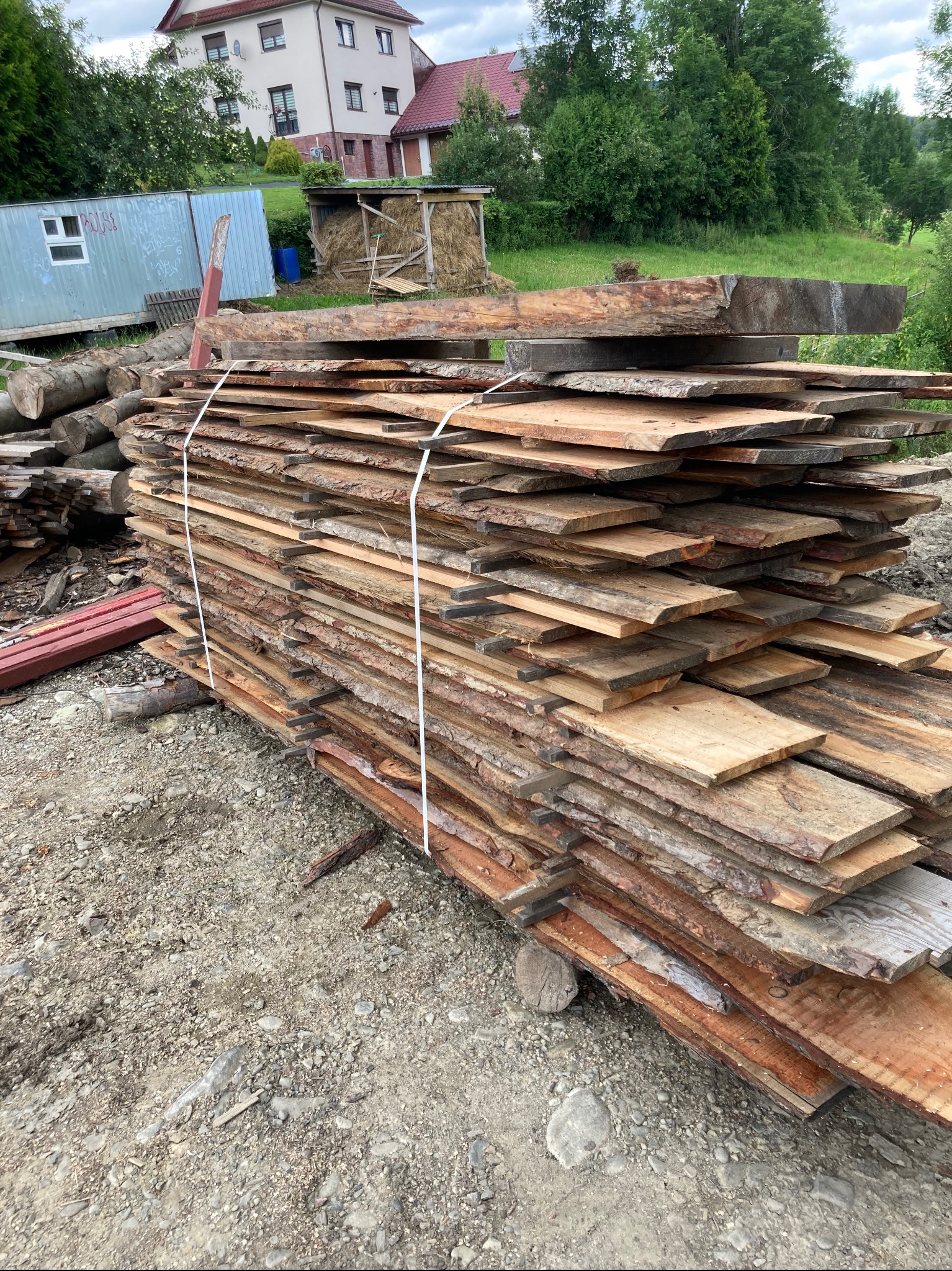 Stos desek z korą, przewiązanych białym sznurkiem, ułożonych na nierównym podłożu z kamieni, w tle budynek mieszkalny i zieleń.