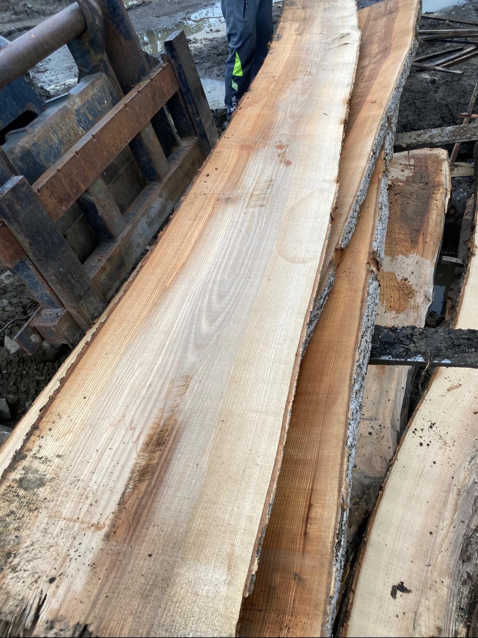 Stos desek z surowego drewna z korą, ułożonych obok metalowego osprzętu ciężkiego sprzętu na zewnątrz, widoczny fragment ubrania osoby w tle.