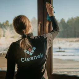 Kobieta w czarnej koszulce z logo firmy 'Cleanek' czyści ramę okienną niebieską ściereczką, widok z tyłu, na zewnątrz krajobraz z drzewami i resztkami śniegu.