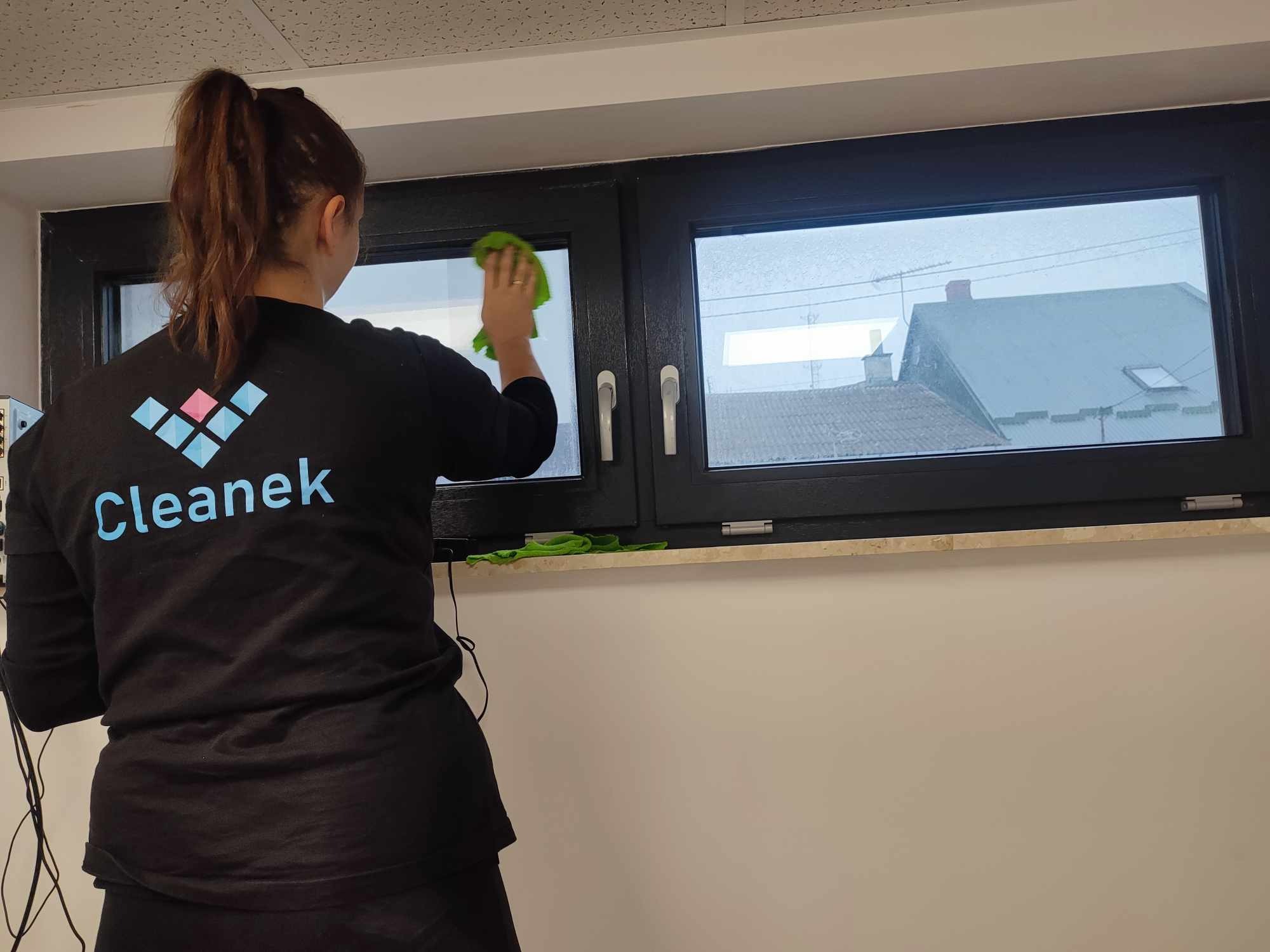 Pracownica firmy Cleank czyści okno szmatką, widok zza pleców, na zewnątrz budynki.