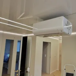 Biała klimatyzacja typu split zamontowana pod sufitem w nowoczesnym wnętrzu z oświetleniem LED, widok z dołu. Na ścianie lustrzane panele.