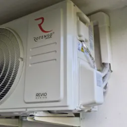 Biały klimatyzator Rotenso Revio Series zamontowany na zewnątrz budynku, widoczne rury odprowadzające, ściana z oknem w tle.